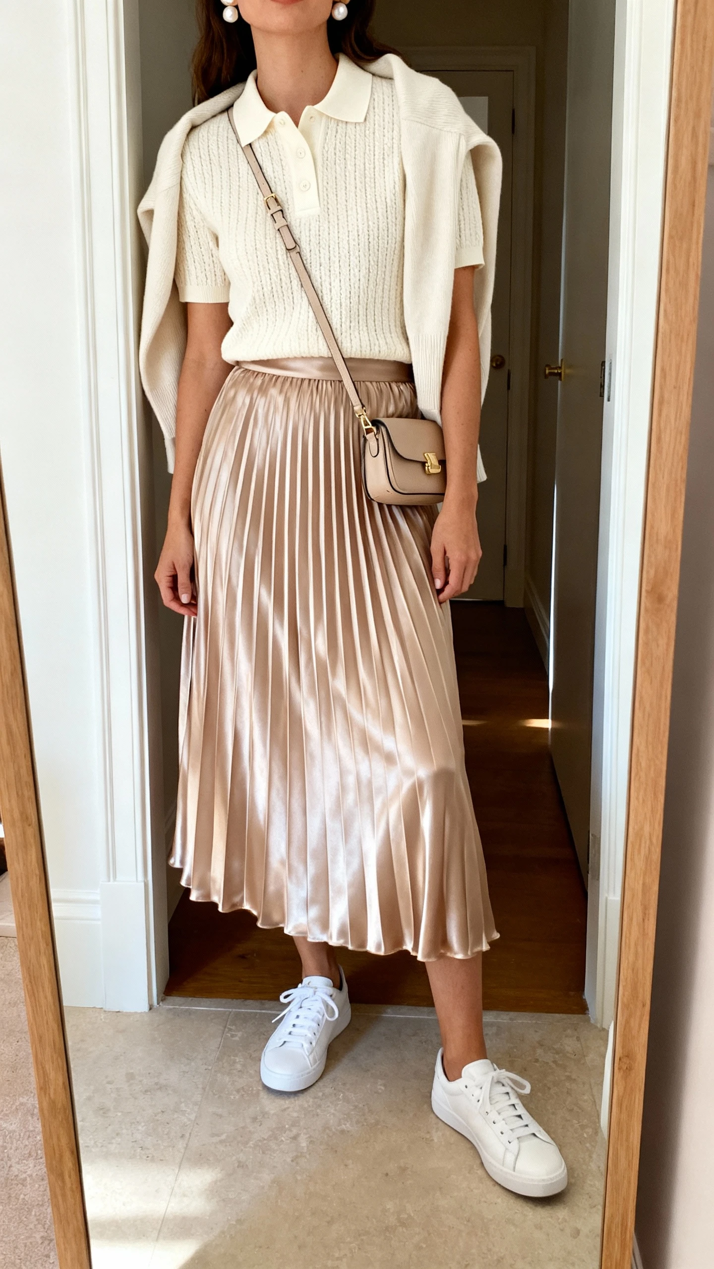 iPhone mirror selfie of a woman in a cream knit polo shirt, midi satin pleated skirt, white sneakers, small satchel crossbody, pearl studs, and a lightweight cardigan draped over the shoulders; face not visible, cropped at neck, entryway full-length mirror, soft indoor light, iPhone photo quality.