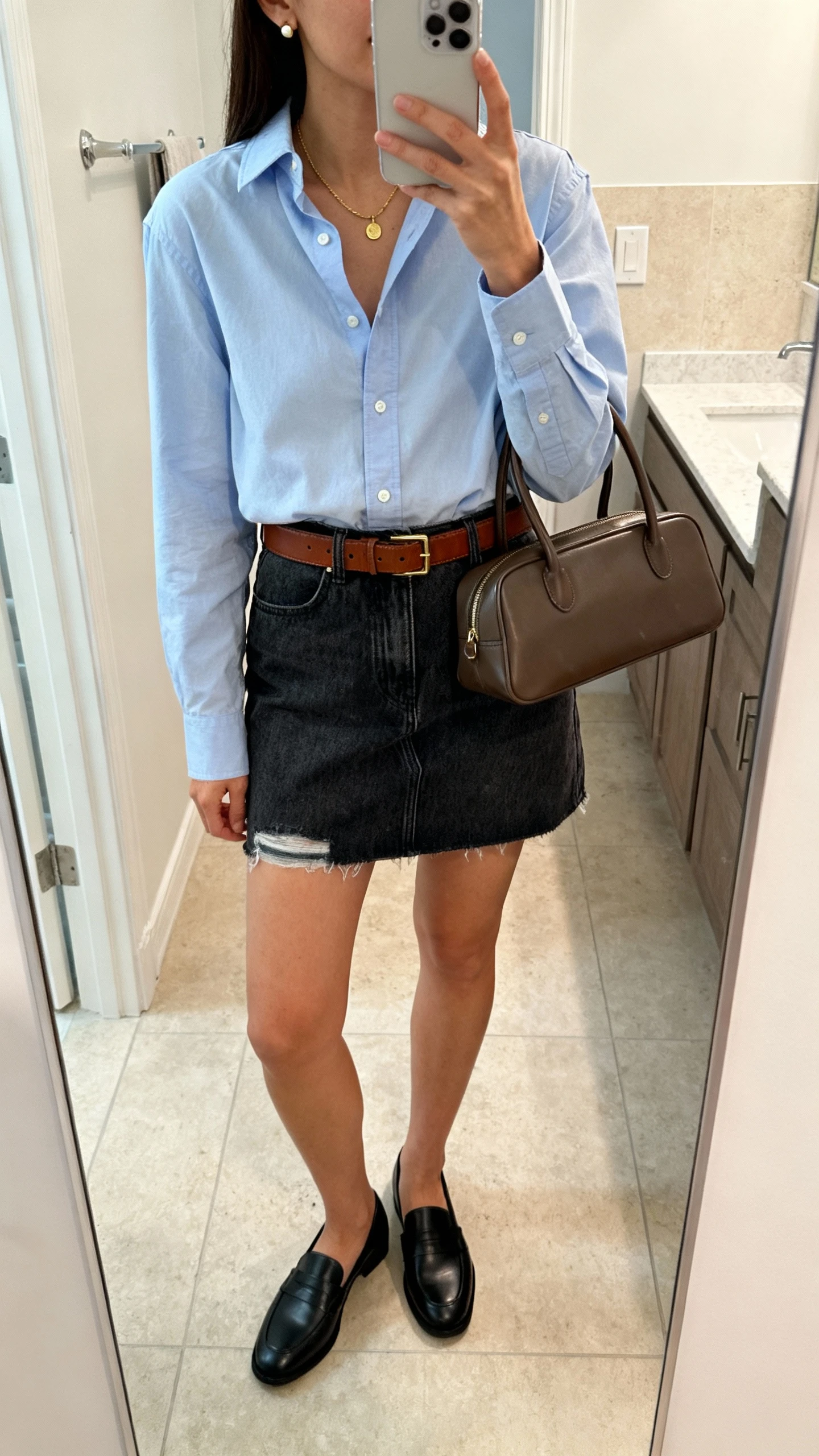 iPhone mirror selfie of a woman in a crisp cotton button-down shirt (light blue), dark-wash denim mini skirt with no distressing, black loafers, chocolate leather belt, structured top-handle bag, delicate gold necklace and stud earrings, face not visible, bathroom mirror, soft indoor lighting, iPhone photo quality.