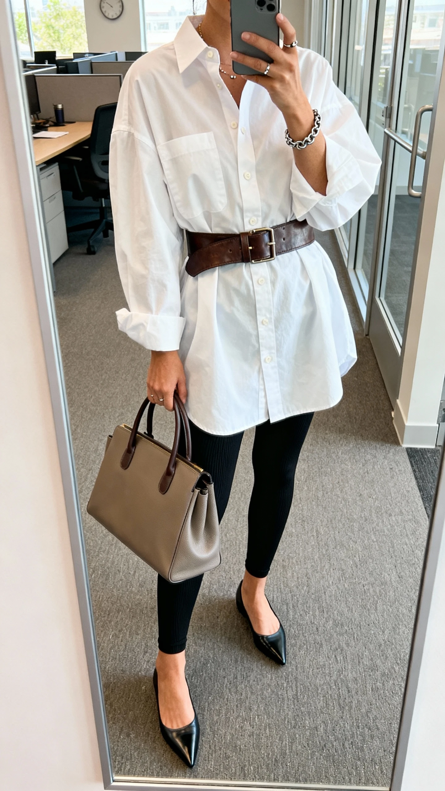 iPhone mirror selfie of a woman in a crisp oversized white button-down shirt with a leather belt over it, ponte ankle leggings in black, pointed-toe flats, a top-handle tote, simple studs and a chain bracelet, face not visible, cropped at shoulders, office entryway mirror, natural daylight, iPhone photo quality.