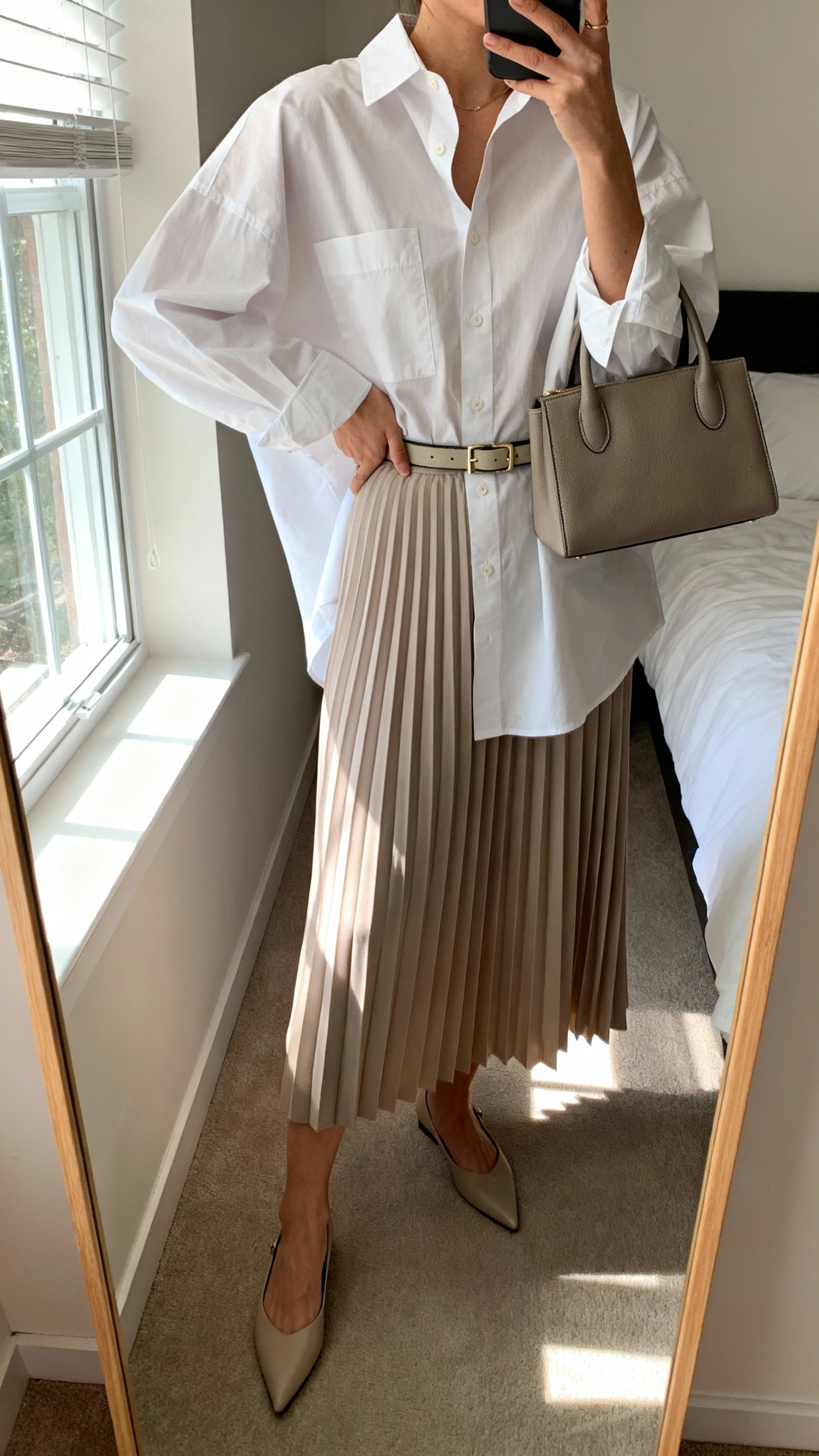 iPhone mirror selfie of a woman in a crisp oversized button-down shirt tucked into a pleated midi skirt, pointed flats, slim belt, structured top-handle bag, face not visible, cropped at neck, bedroom mirror with window light, iPhone photo quality.