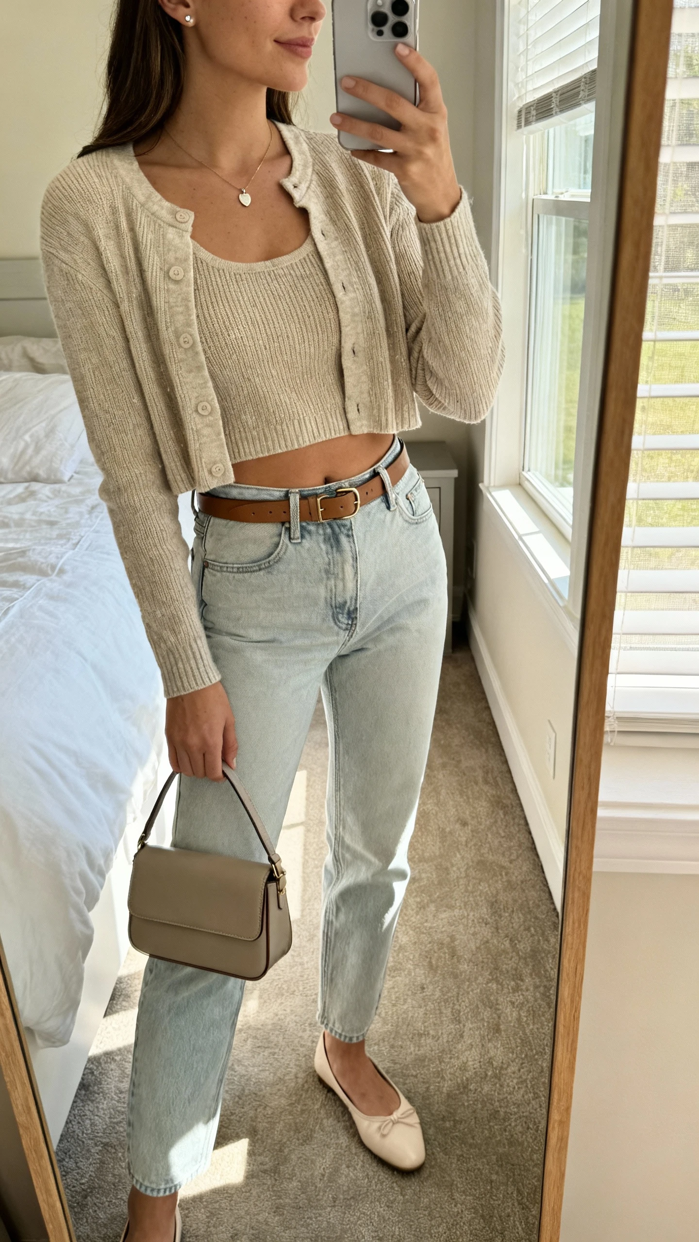 iPhone mirror selfie of a woman in a cropped cardigan with matching knit tank, light-wash mom jeans, ballet flats, a small pendant necklace, simple studs, a slim leather belt, and a structured mini tote, face not visible, side angle, bedroom mirror with natural window light, iPhone photo quality.