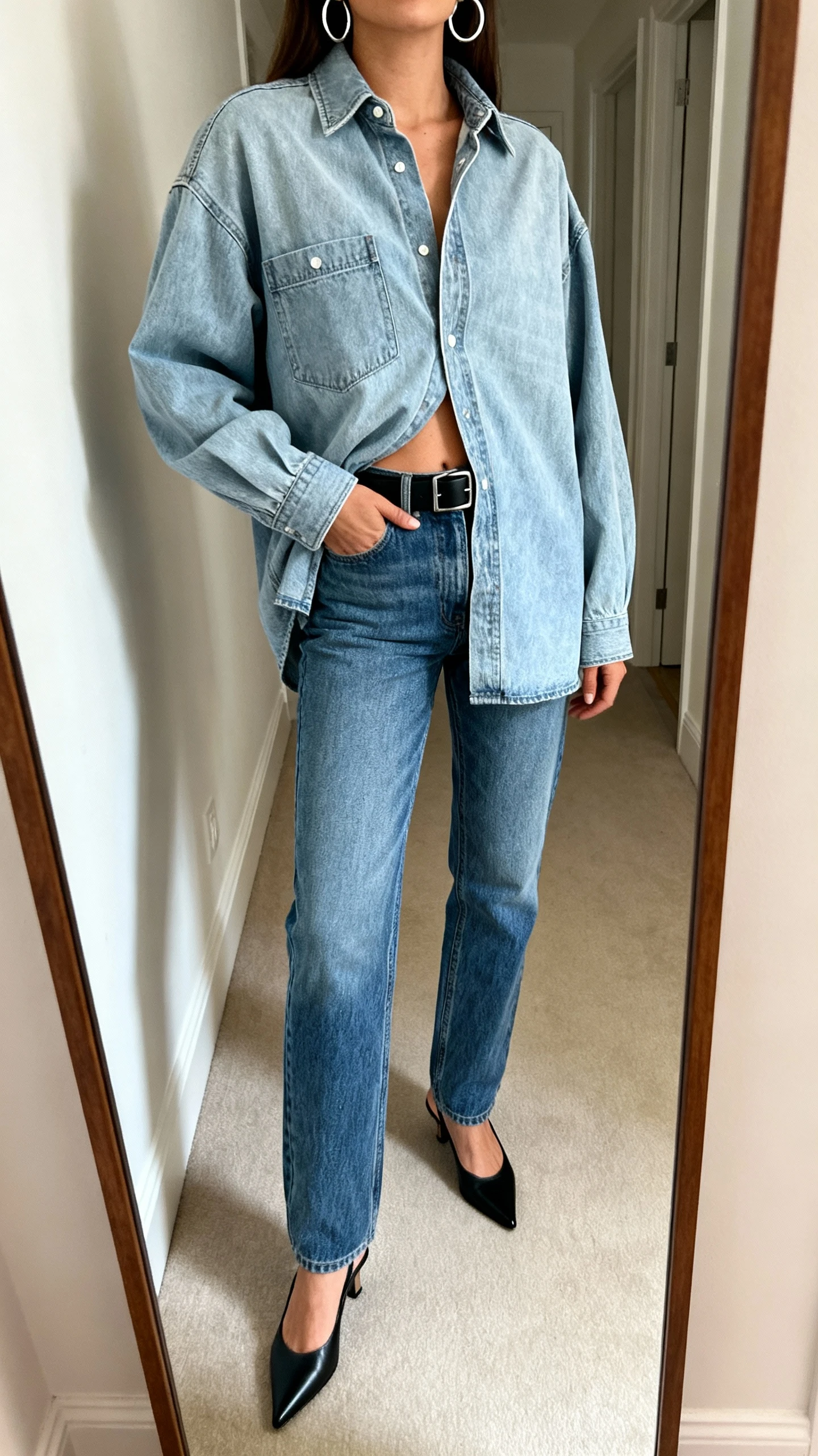 iPhone mirror selfie of a woman in a denim-on-denim look: light-wash oversized denim shirt half-tucked into mid-wash straight-leg jeans, slim black belt, silver hoops, pointed slingback flats, face not visible, cropped at shoulders, hallway full-length mirror, soft indoor light, iPhone photo quality.