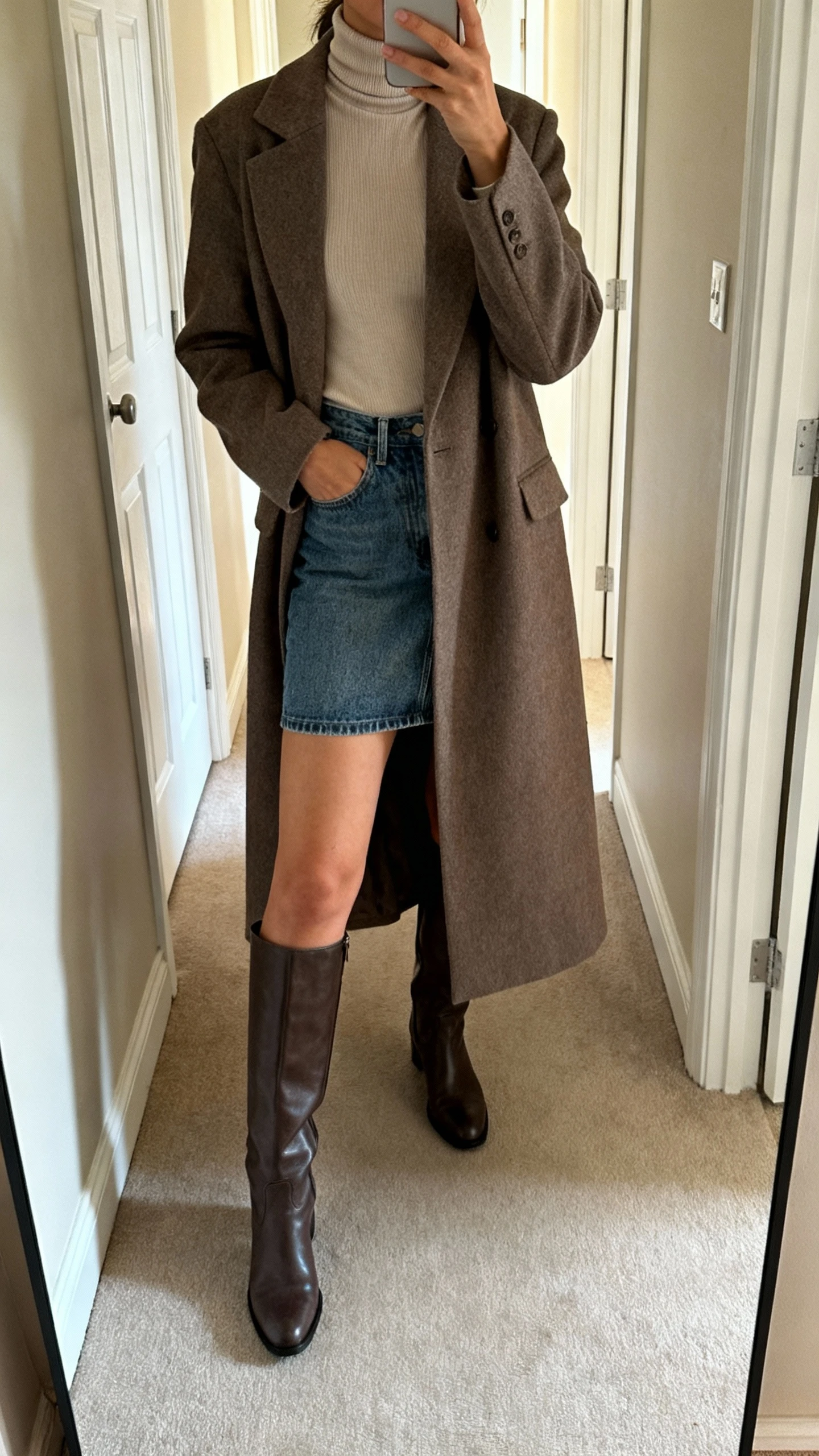 iPhone mirror selfie of a woman in a fitted turtleneck, denim mini skirt, structured longline coat, and knee-high boots, face not visible, cropped at shoulders, hallway mirror, soft morning light, iPhone photo quality.