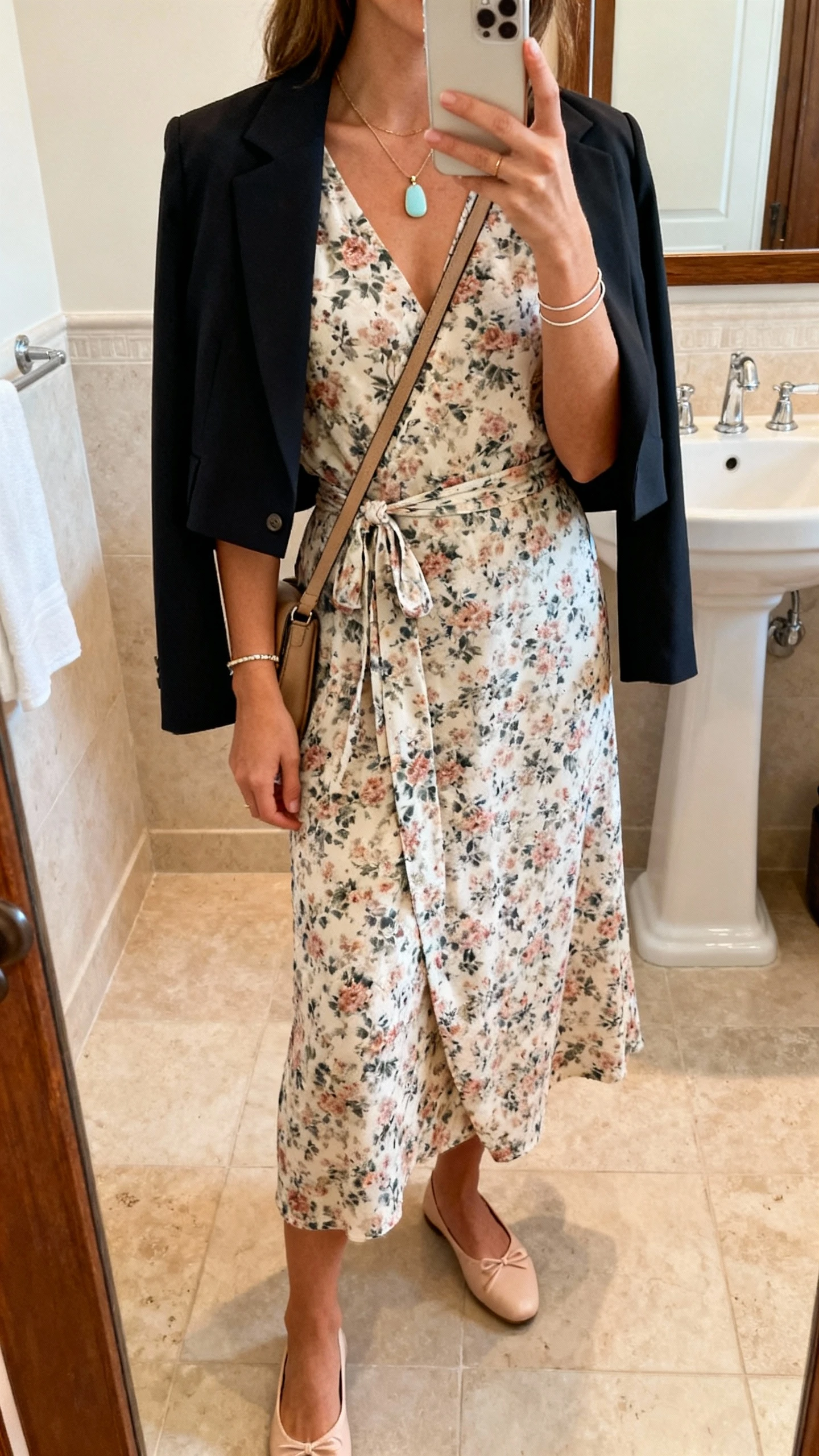 iPhone mirror selfie of a woman in a floral midi wrap dress with a cropped blazer draped over shoulders, ballet flats, light pendant necklace, thin bracelets, and a crossbody bag; face not visible, bathroom mirror, warm indoor lighting, iPhone photo quality.