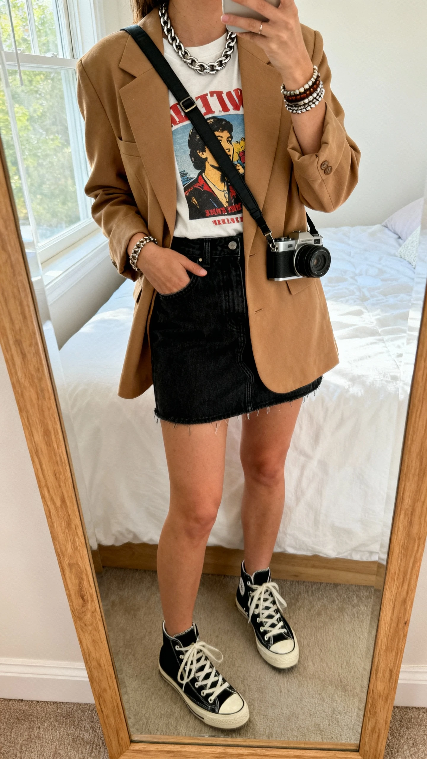 iPhone mirror selfie of a woman in a graphic T-shirt under a camel boyfriend blazer, black denim mini skirt, high-top sneakers, crossbody camera bag, chunky chain necklace and stacked bracelets, face not visible, cropped at shoulders, bedroom mirror, natural window light, iPhone photo quality.