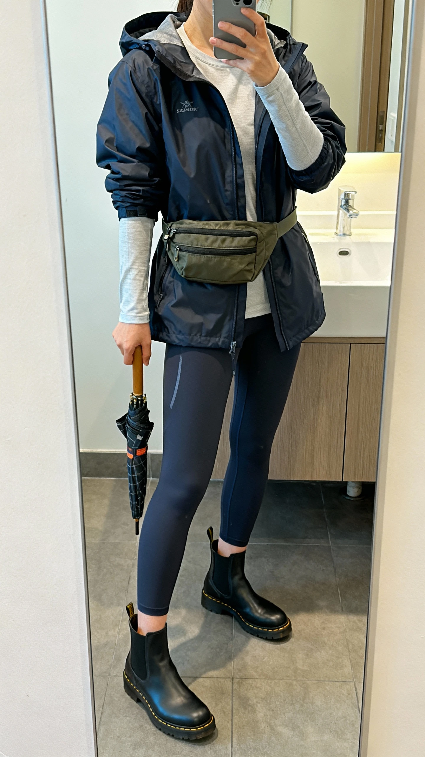 iPhone mirror selfie of a woman in a hooded water-resistant parka, quick-dry leggings, rubberized Chelsea boots, light long-sleeve base layer, nylon crossbody belt bag, compact umbrella in hand, face not visible, cropped at neck, bathroom mirror, indoor lighting, iPhone photo quality.