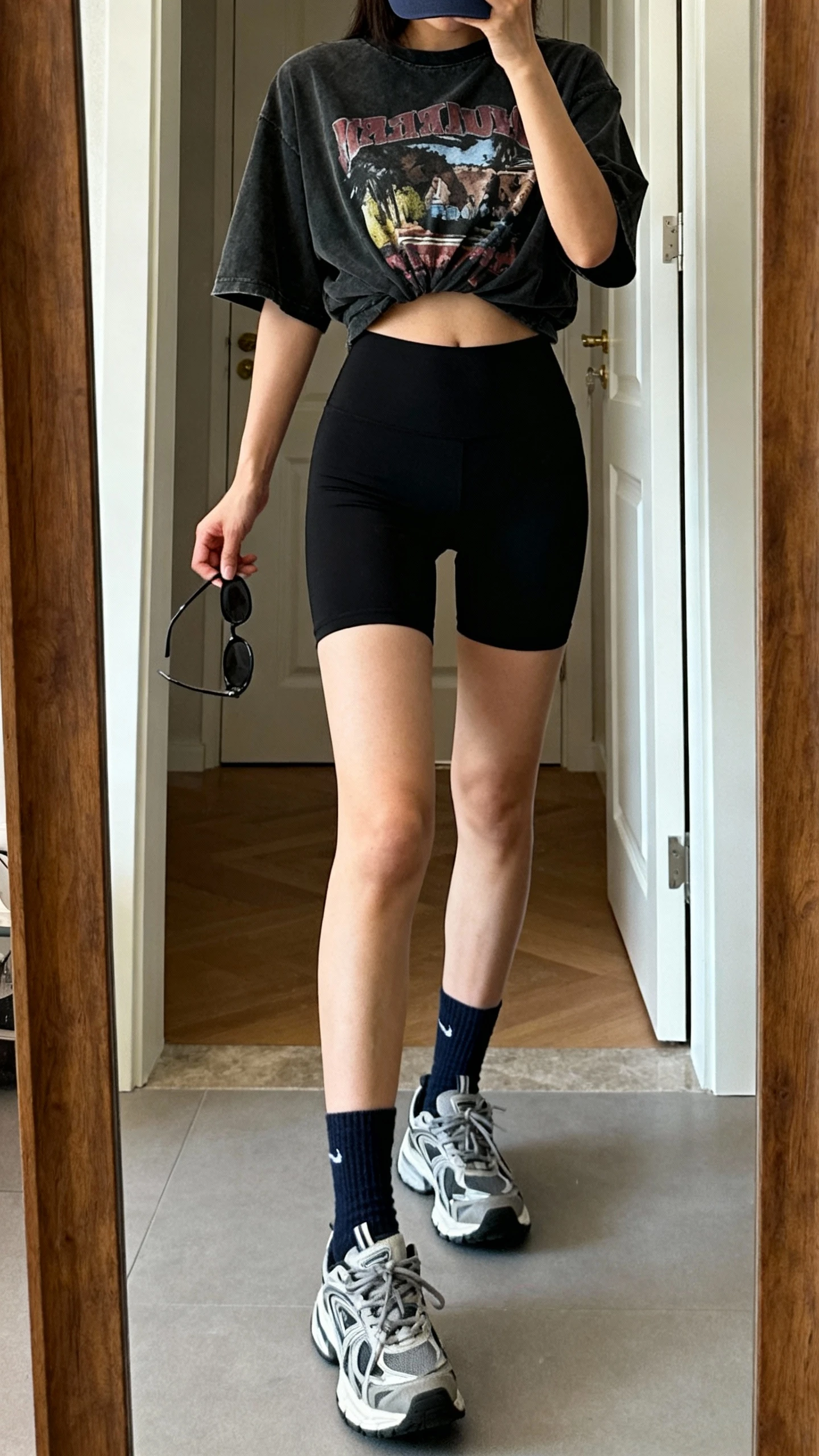 iPhone mirror selfie of a woman in a hot-girl walk fit: black biker shorts, oversized graphic tee half-tucked, crew socks, chunky sneakers, baseball cap, and simple sunglasses in hand, face not visible, cropped at shoulders, entryway full-length mirror, natural daylight, iPhone photo quality.