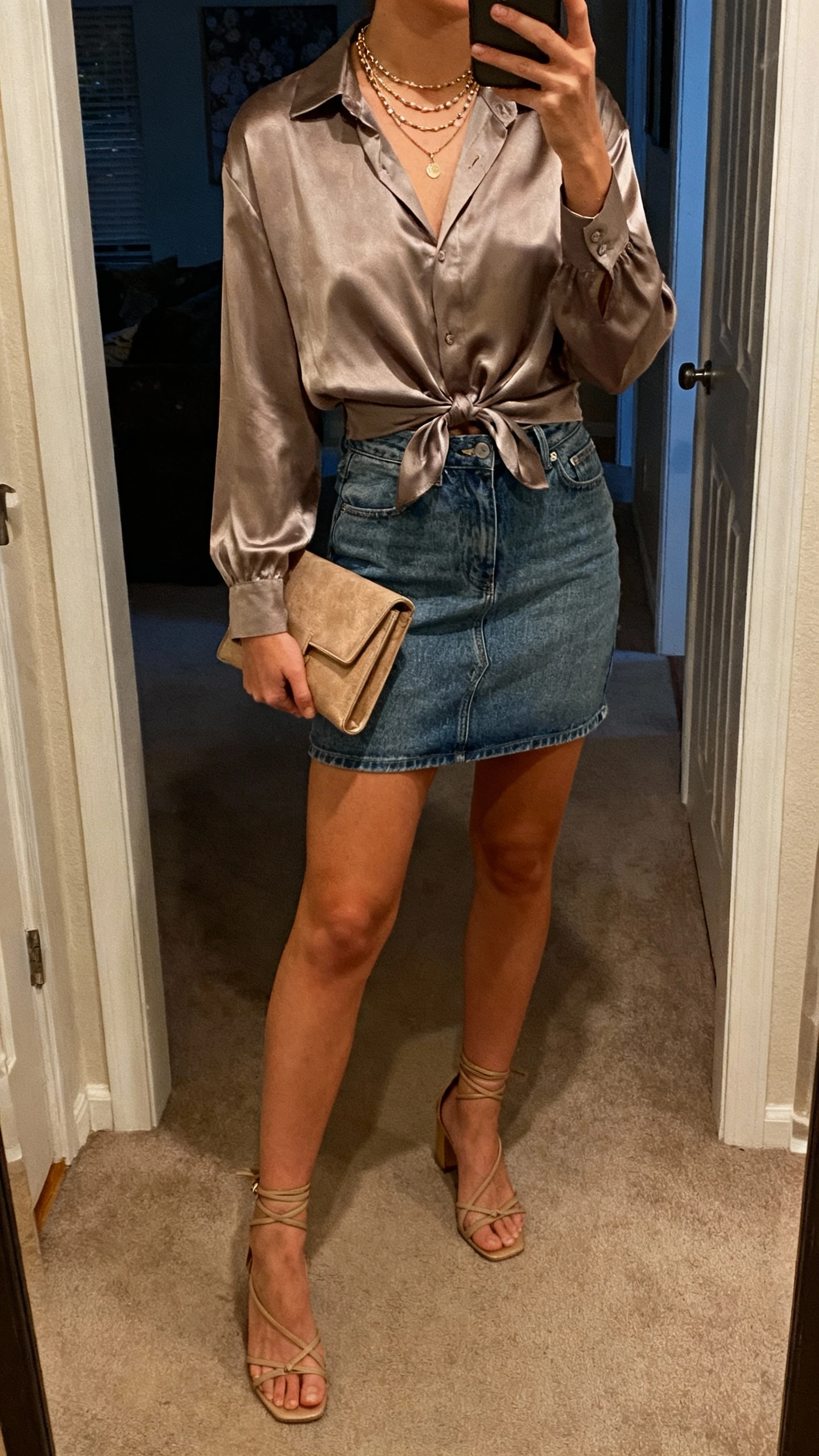 iPhone mirror selfie of a woman in a jeans mini skirt with a silky blouse tucked in and strappy heels, clutch bag, layered necklaces, face not visible, cropped at neck, entryway mirror, evening indoor light, iPhone photo quality.