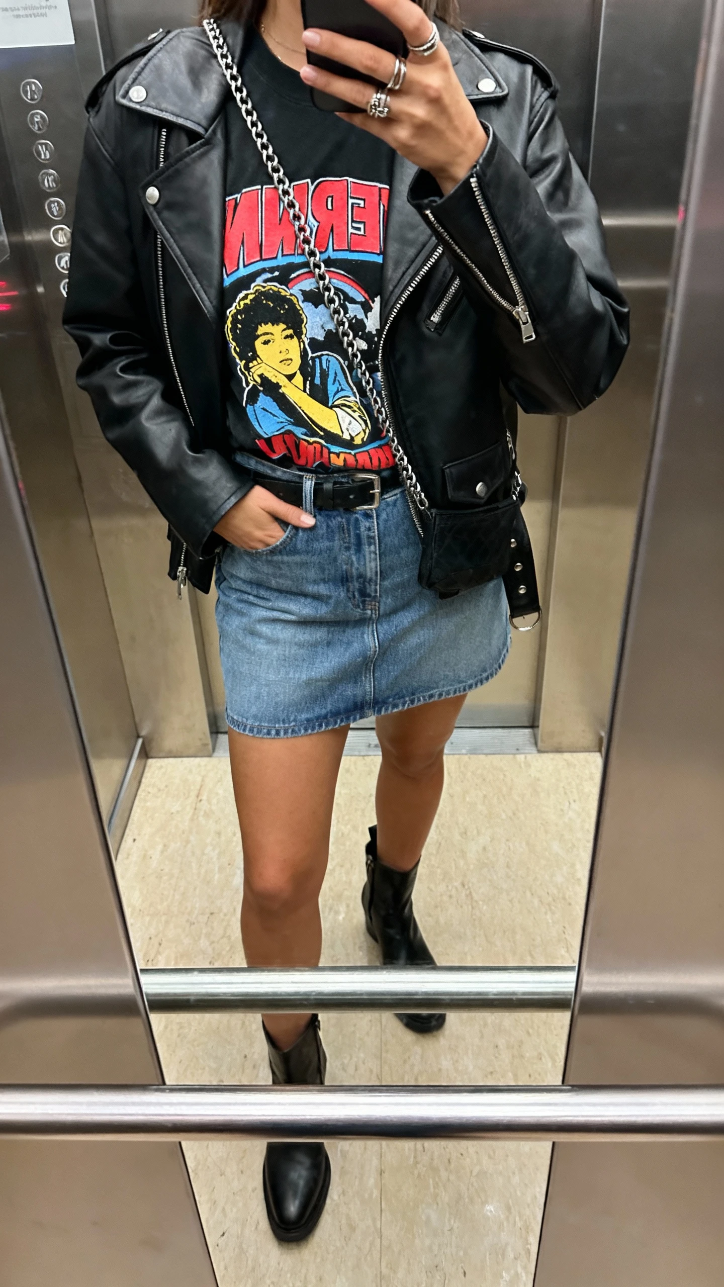 iPhone mirror selfie of a woman in a jeans mini skirt with a bold graphic tee, black moto jacket, and ankle boots, chain crossbody, rings, face not visible, cropped at shoulders, elevator mirror, indoor lighting, iPhone photo quality.