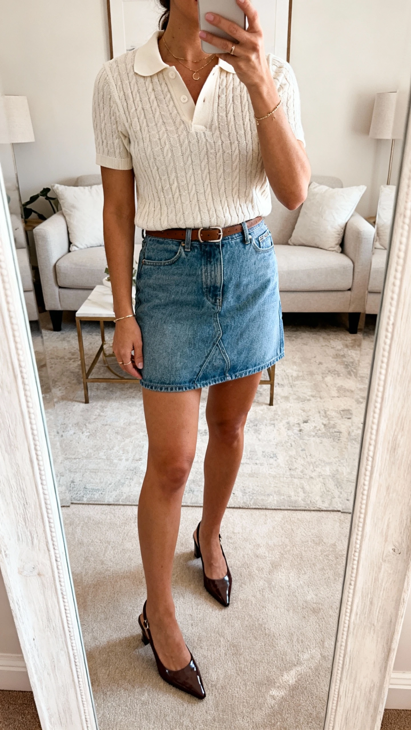 iPhone mirror selfie of a woman in a knit polo tucked into a denim mini skirt with sleek slingback shoes and minimal jewelry, face not visible, cropped at neck, living room mirror, soft morning light, iPhone photo quality.
