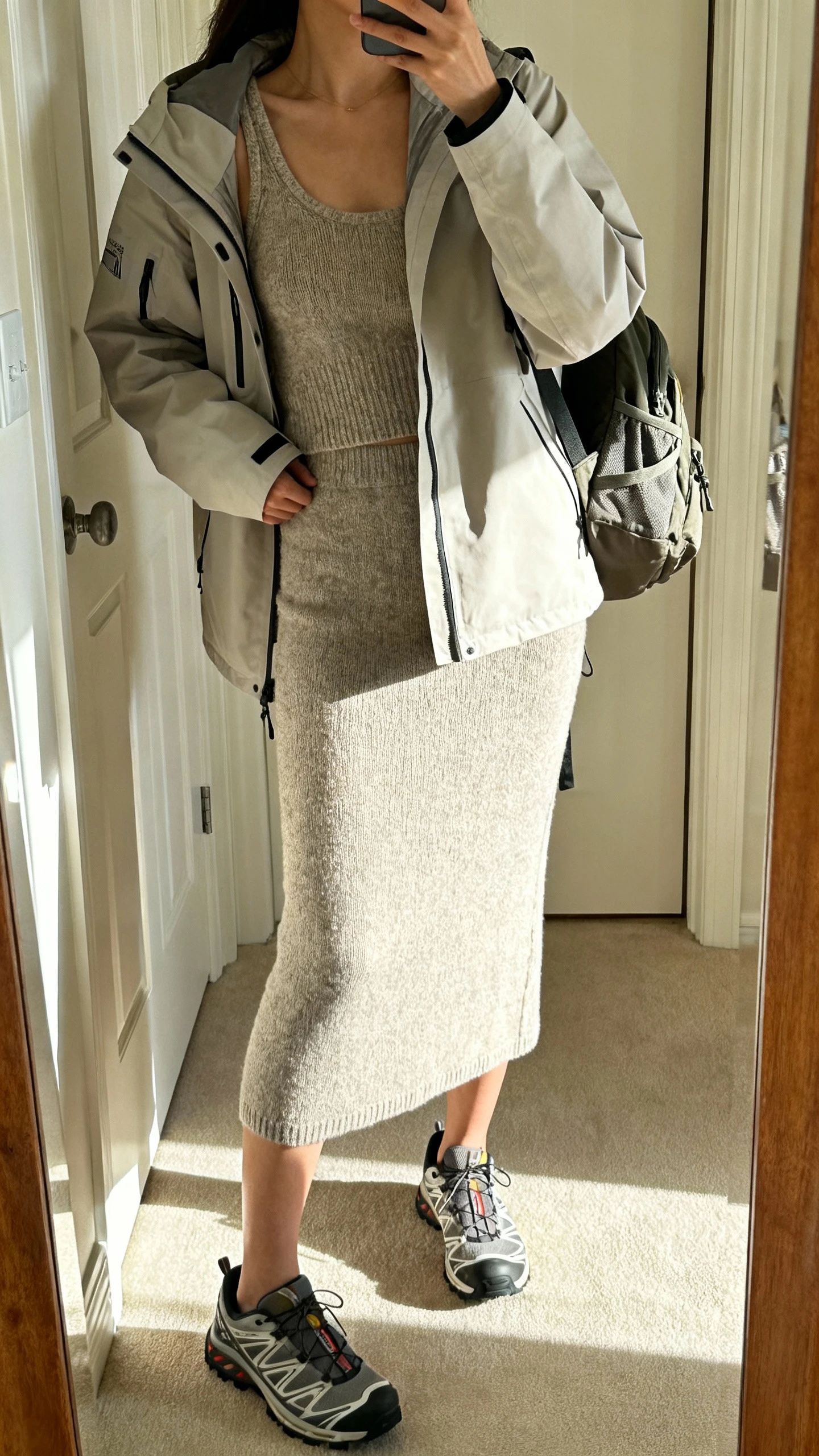 iPhone mirror selfie of a woman in a lightweight parka over a knit tank-and-skirt set with trail-inspired sneakers and a small backpack, face not visible, cropped at neck, entryway mirror, morning daylight, iPhone photo quality.