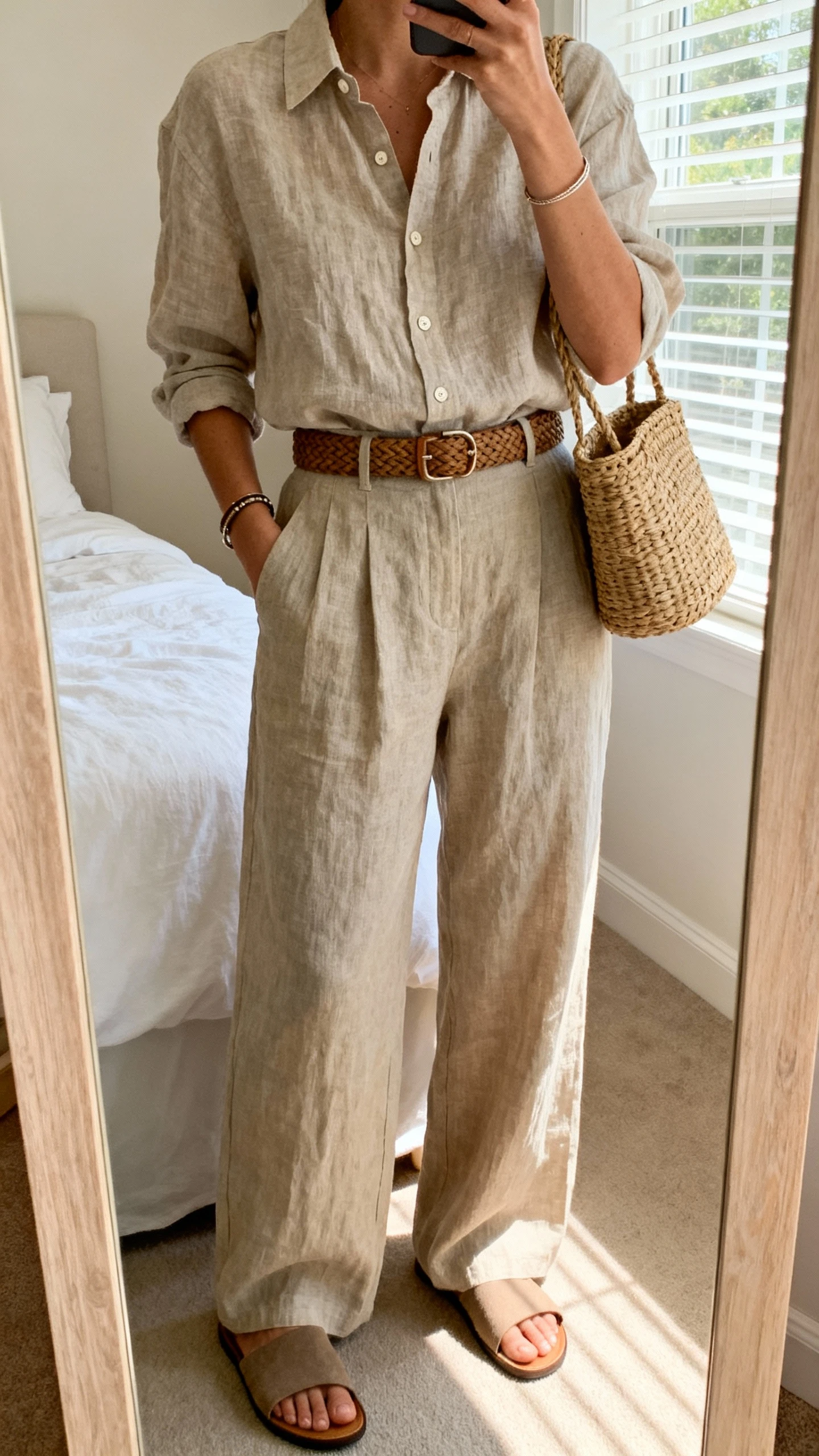 iPhone mirror selfie of a woman in a linen button-up shirt tucked into linen wide-leg trousers, flat slide sandals, woven belt, rattan bag, simple bracelets, face not visible, cropped at shoulders, bedroom mirror, soft afternoon window light, iPhone photo quality.