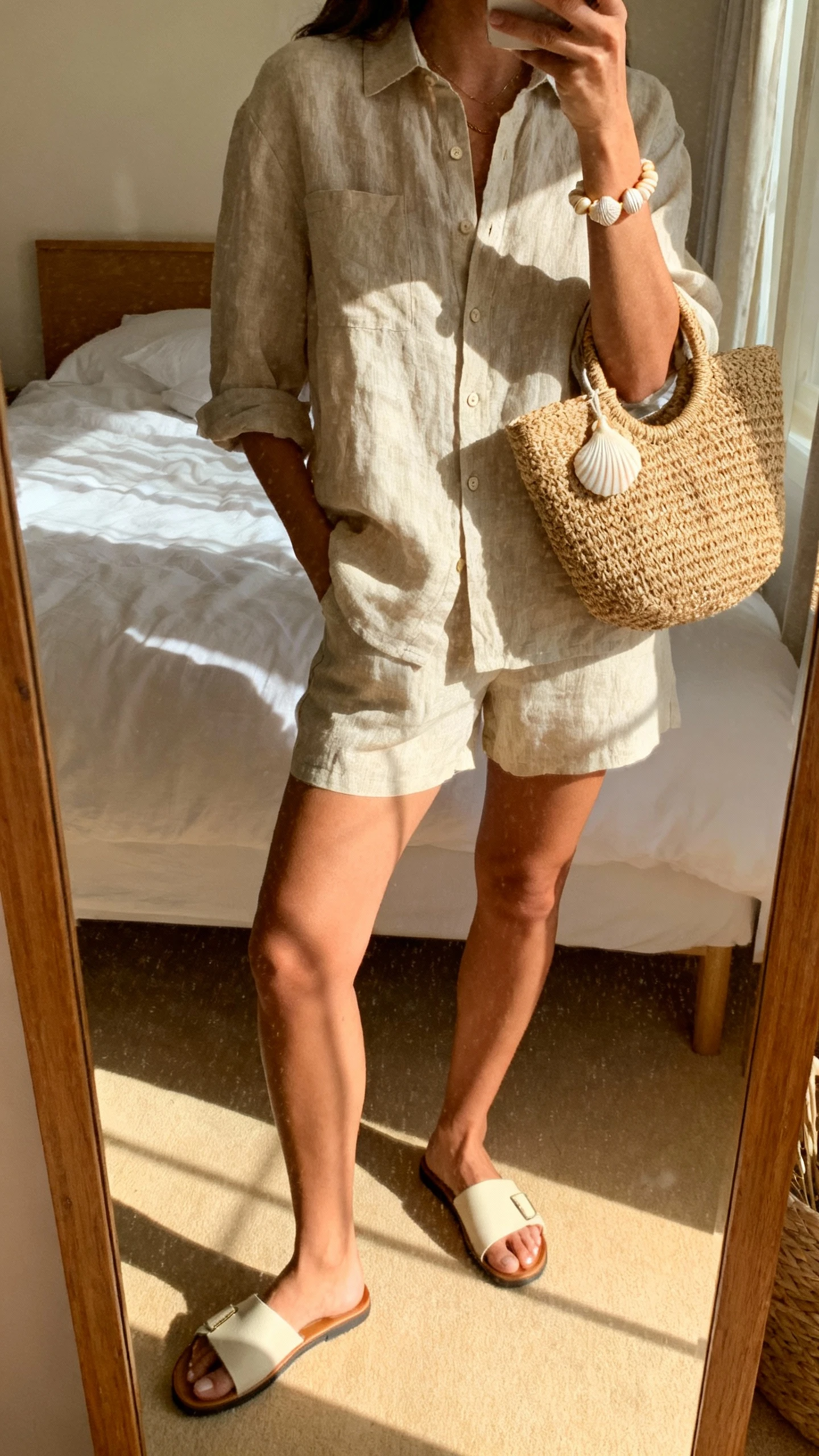 iPhone mirror selfie of a woman in a linen co-ord set (button shirt and matching shorts) with slide sandals, straw tote, and shell bracelet, face not visible, cropped at shoulders, sunlit bedroom mirror, iPhone photo quality.