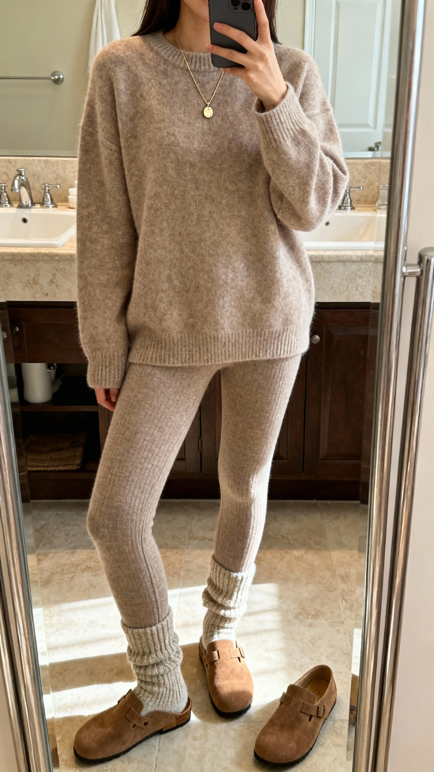 iPhone mirror selfie of a woman in a matching cashmere-effect sweater set: soft crewneck sweater with coordinating knit leggings, cozy socks, delicate pendant necklace, suede loafers beside her, face not visible, bathroom full-length mirror, warm indoor lighting, iPhone photo quality.