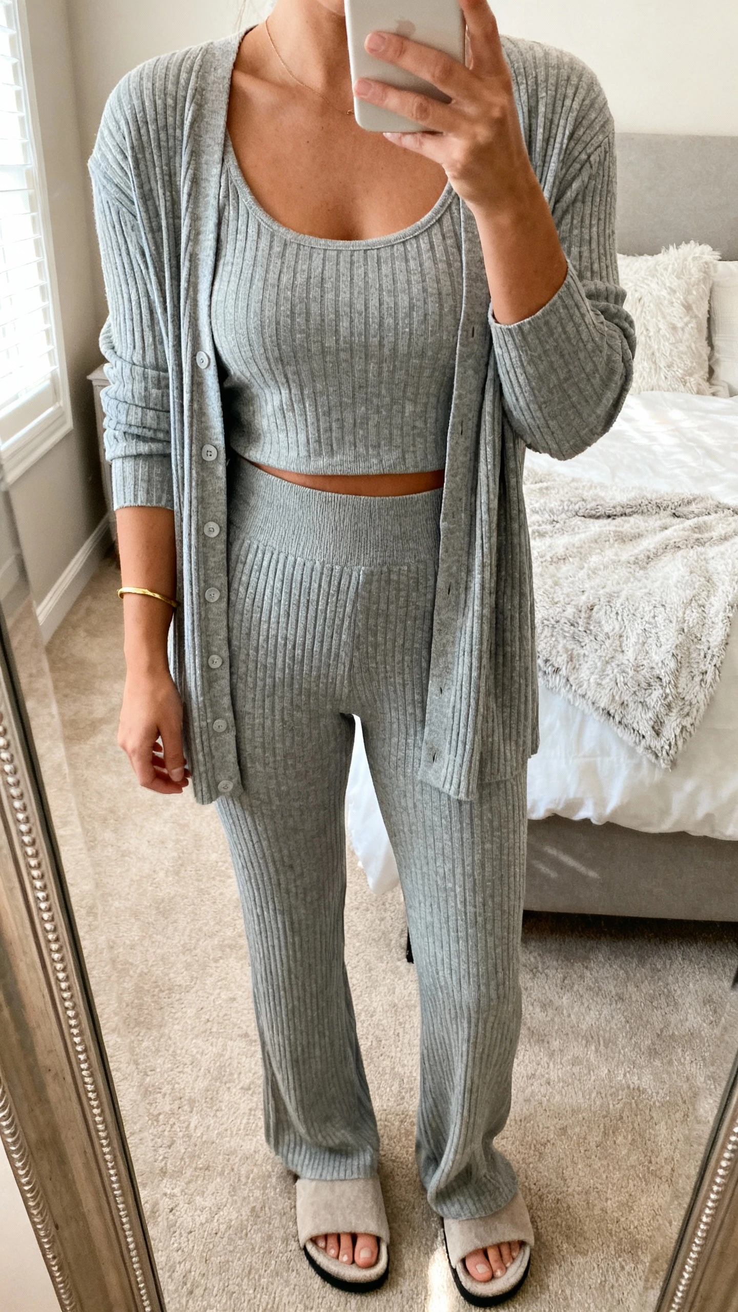 iPhone mirror selfie of a woman in a matching knit set: ribbed cardigan and high-rise knit pants in heather gray, simple tank underneath, thin gold bracelet, cushy slide sandals, face not visible, cropped at neck, cozy bedroom mirror, warm indoor light, iPhone photo quality.