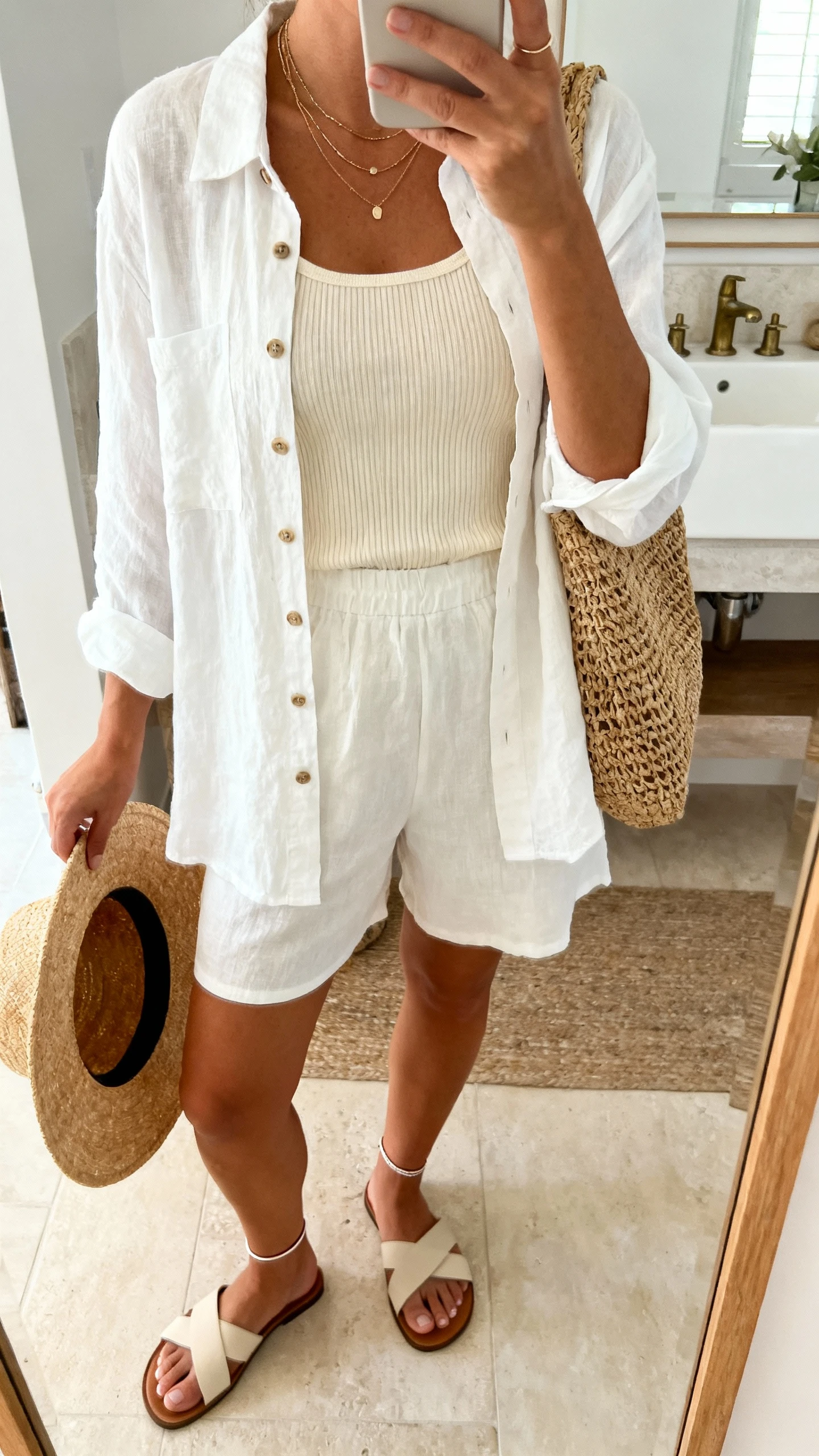 iPhone mirror selfie of a woman in a matching white linen button-down and shorts, cream ribbed tank, minimal slide sandals, woven tote, layered delicate necklaces and thin anklet, wide-brim straw hat in hand; face not visible, cropped at shoulders, bathroom mirror, soft morning light, iPhone photo quality.