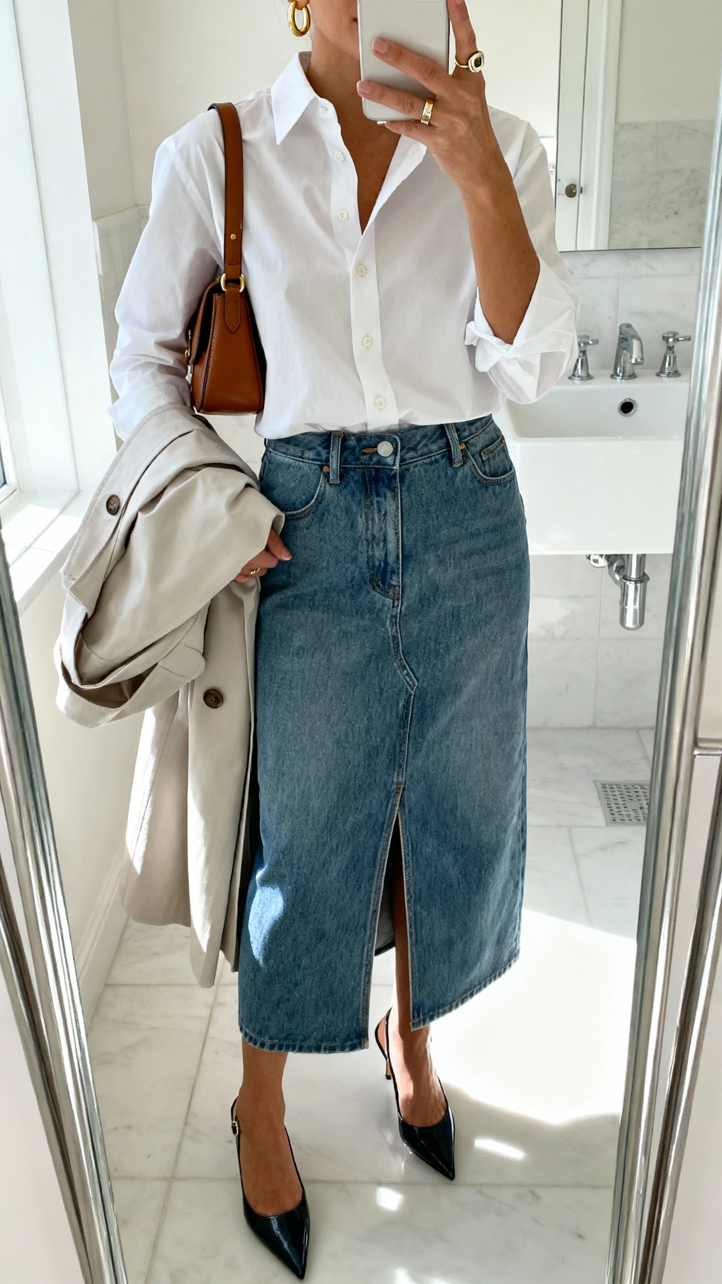 iPhone mirror selfie of a woman in a medium-wash denim midi skirt with a front slit, crisp white button-up shirt, pointed kitten-heel slingbacks, leather shoulder bag, gold huggie hoops, signet ring, and a light trench coat over her arm, face not visible, cropped at neck, bathroom mirror with bright natural light, iPhone photo quality.