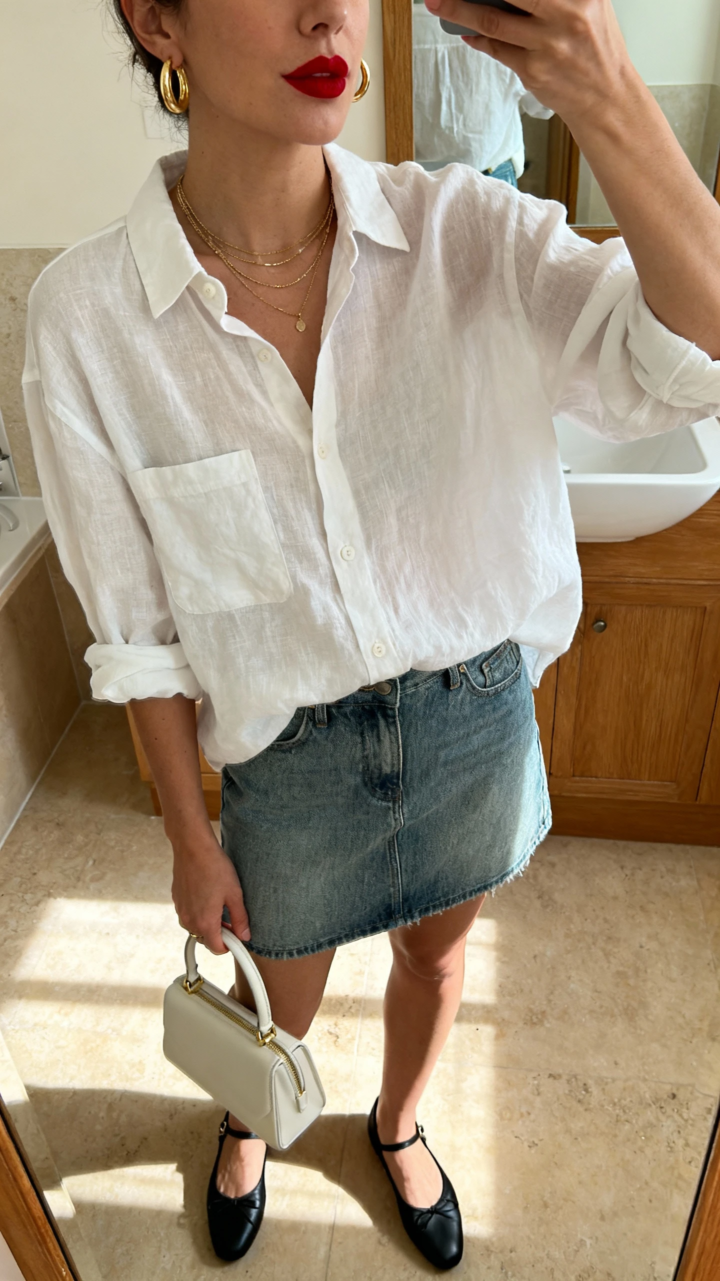 iPhone mirror selfie of a woman in a mid-wash denim mini skirt, slightly oversized white linen button-down with sleeves rolled, black ballet flats, a mini top-handle bag, gold hoop earrings, delicate layered necklaces, and classic red lipstick; face not visible (cropped at nose), bathroom mirror, morning light, iPhone photo quality.