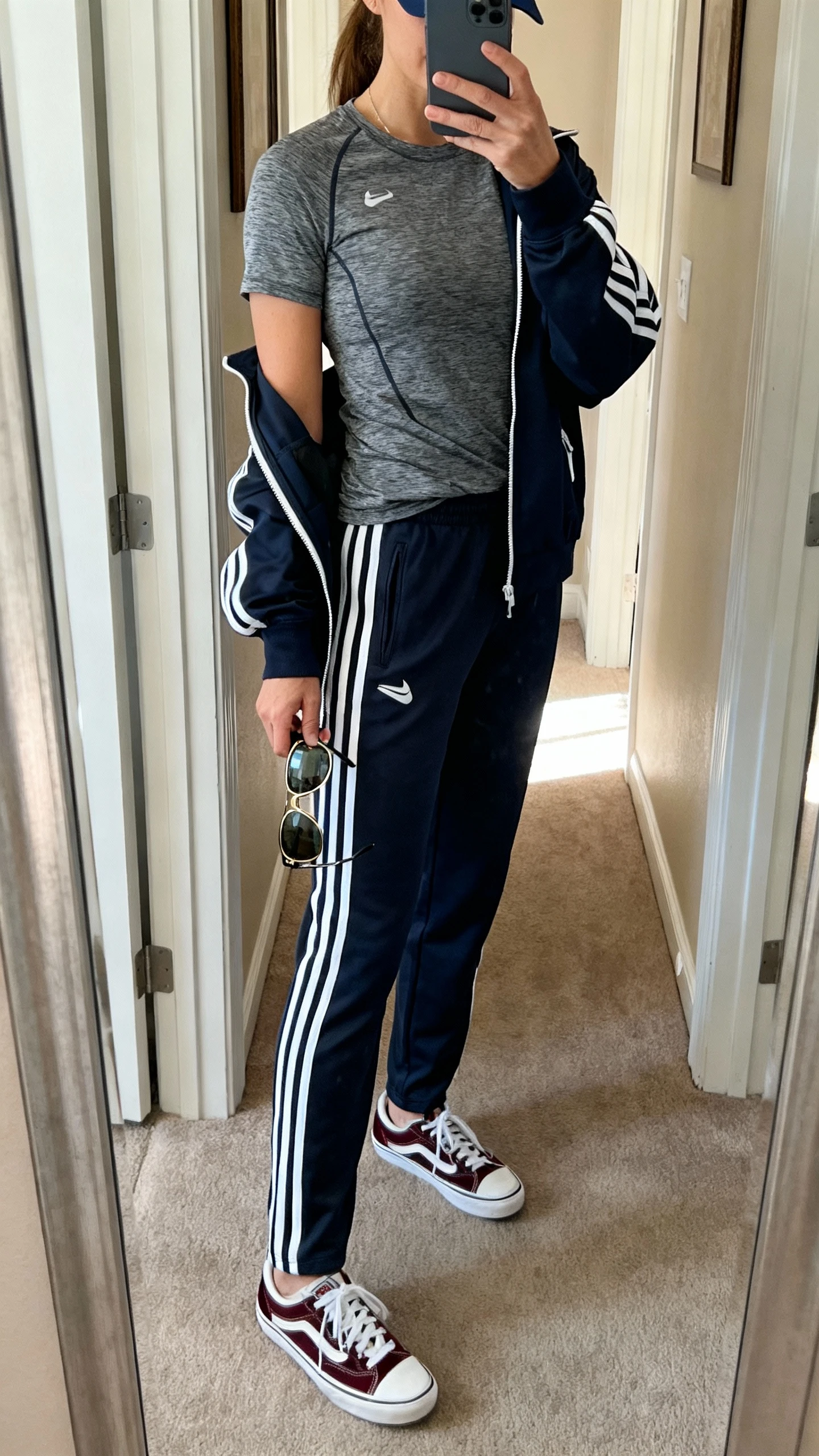 iPhone mirror selfie of a woman in a moisture-wicking tech tee, striped track pants, retro-inspired trainers, zip-up track jacket, sport cap and aviator-style sunglasses in hand, face not visible, cropped at neck, entryway full-length mirror with afternoon daylight, iPhone photo quality