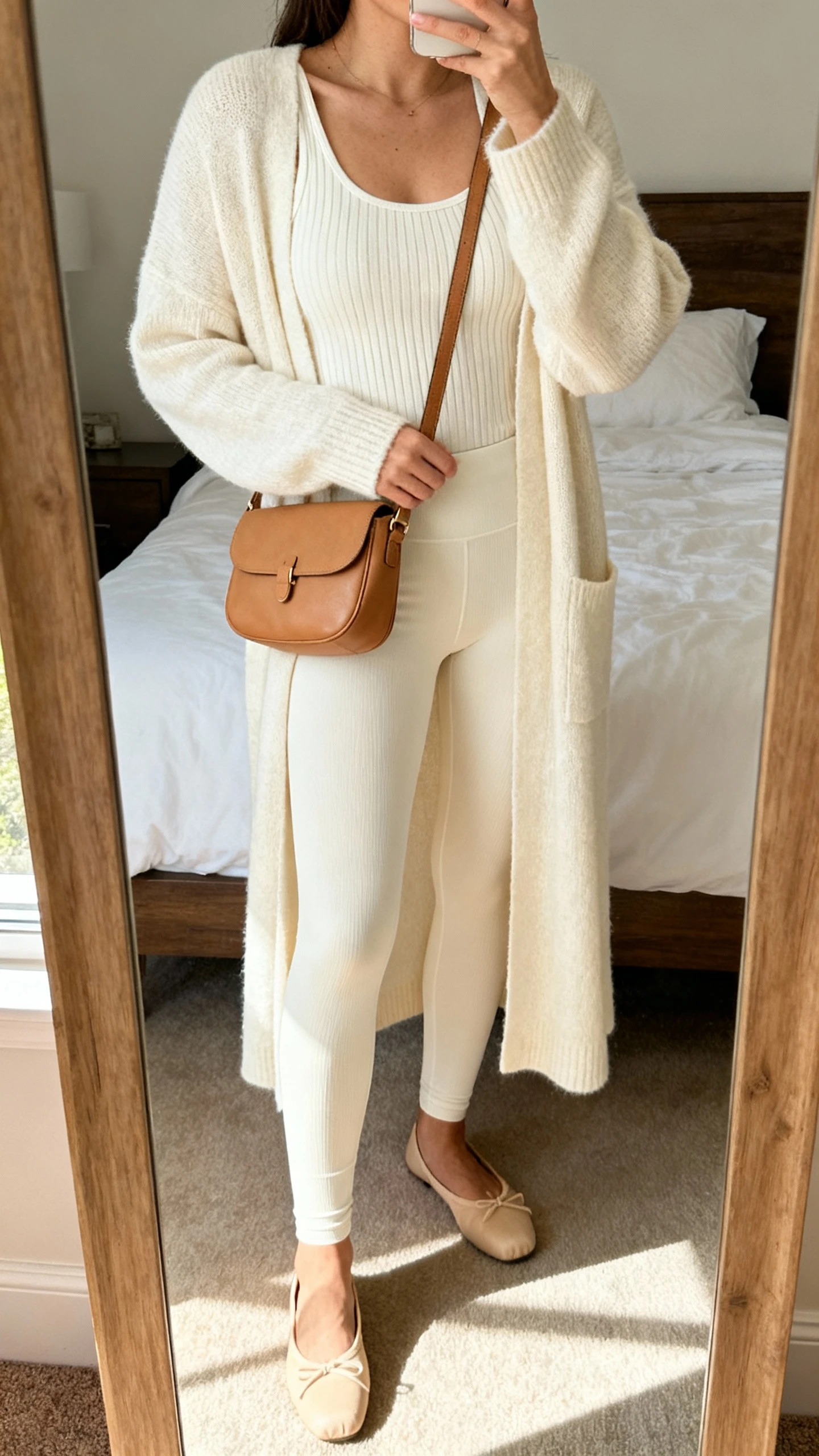 iPhone mirror selfie of a woman in a monochrome set: cream long cardigan, cream leggings, cream ribbed tank, tan crossbody bag, and beige ballet flats, face not visible, cropped at neck, bedroom mirror, soft afternoon light, iPhone photo quality.