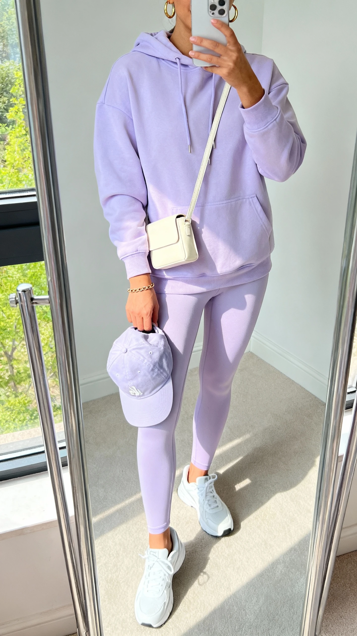 iPhone mirror selfie of a woman in a pastel lavender hoodie with matching leggings, clean white sneakers, mini cream crossbody, simple gold hoops and a slim chain bracelet, optional baseball cap in hand, face not visible, cropped at neck, full-length mirror by window, bright daylight, iPhone photo quality.