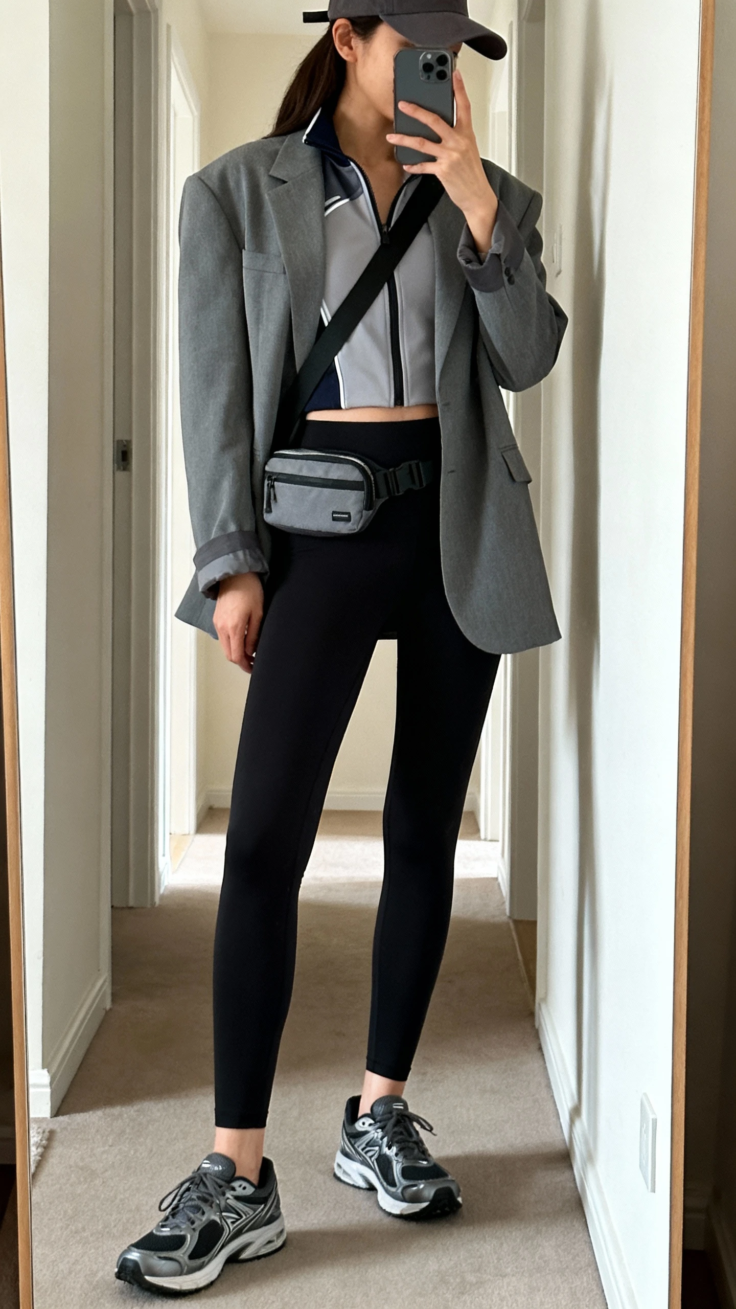 iPhone mirror selfie of a woman in a relaxed gray blazer over a sporty zip-up track top, high-waist black leggings, sleek running sneakers, crossbody belt bag, baseball cap, face not visible, side angle in hallway mirror, indoor natural light, iPhone photo quality.