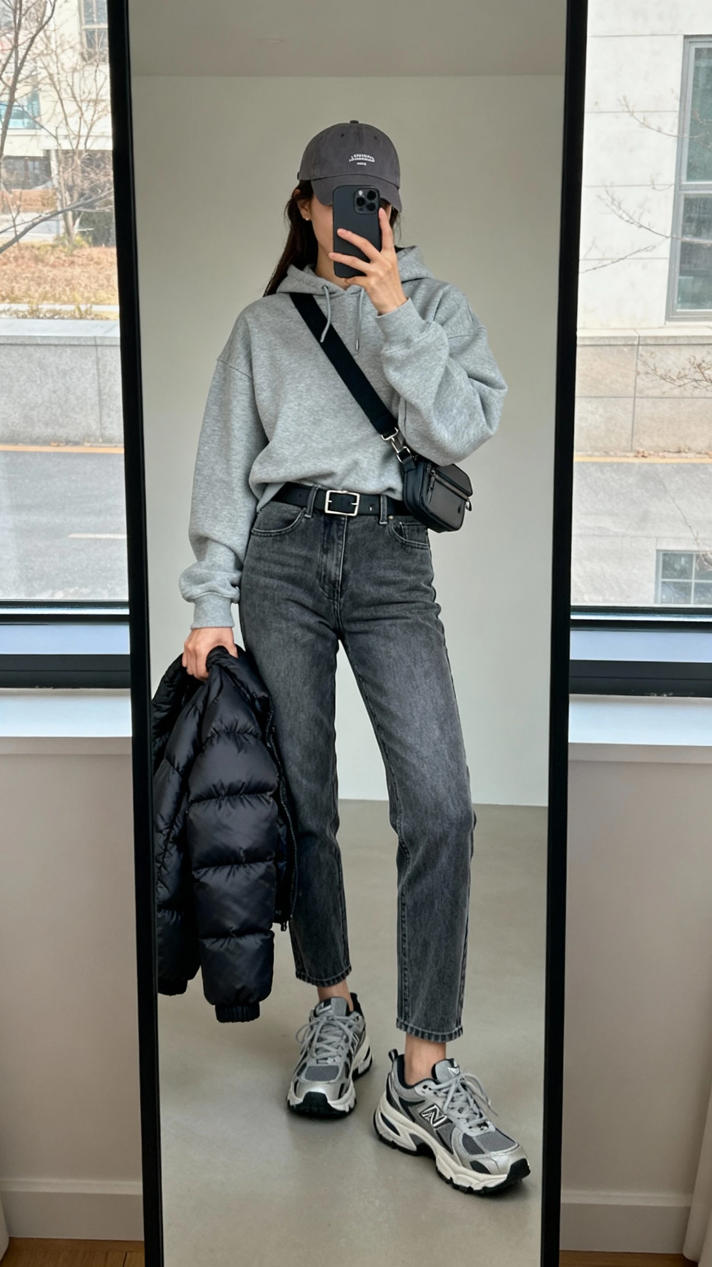 iPhone mirror selfie of a woman in a relaxed grey hoodie, mid-wash tapered mom jeans, chunky sneakers, a baseball cap, and a crossbody belt bag, puffer jacket slung over forearm, face not visible, full-length mirror by window, soft daylight, iPhone photo quality.
