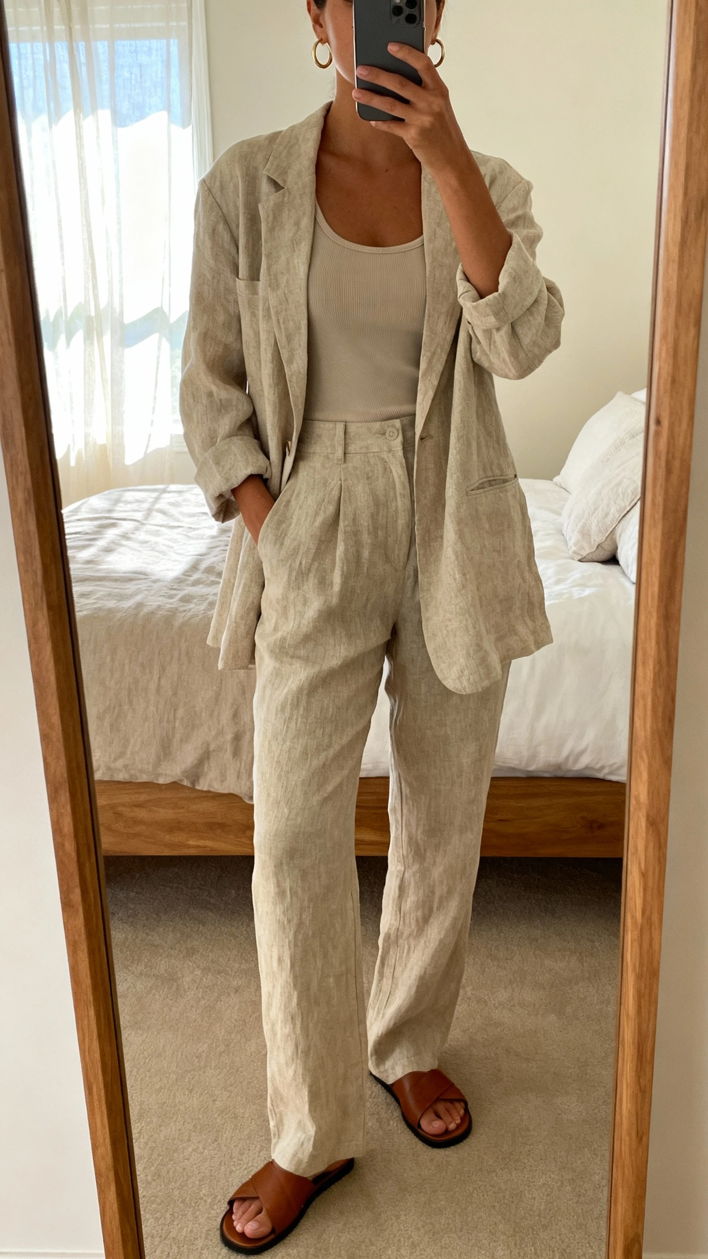 iPhone mirror selfie of a woman in a relaxed linen suit set (unstructured blazer and matching trousers) with a fitted tank and leather slides, simple hoop earrings, face not visible, bedroom full-length mirror, afternoon window light, iPhone photo quality.