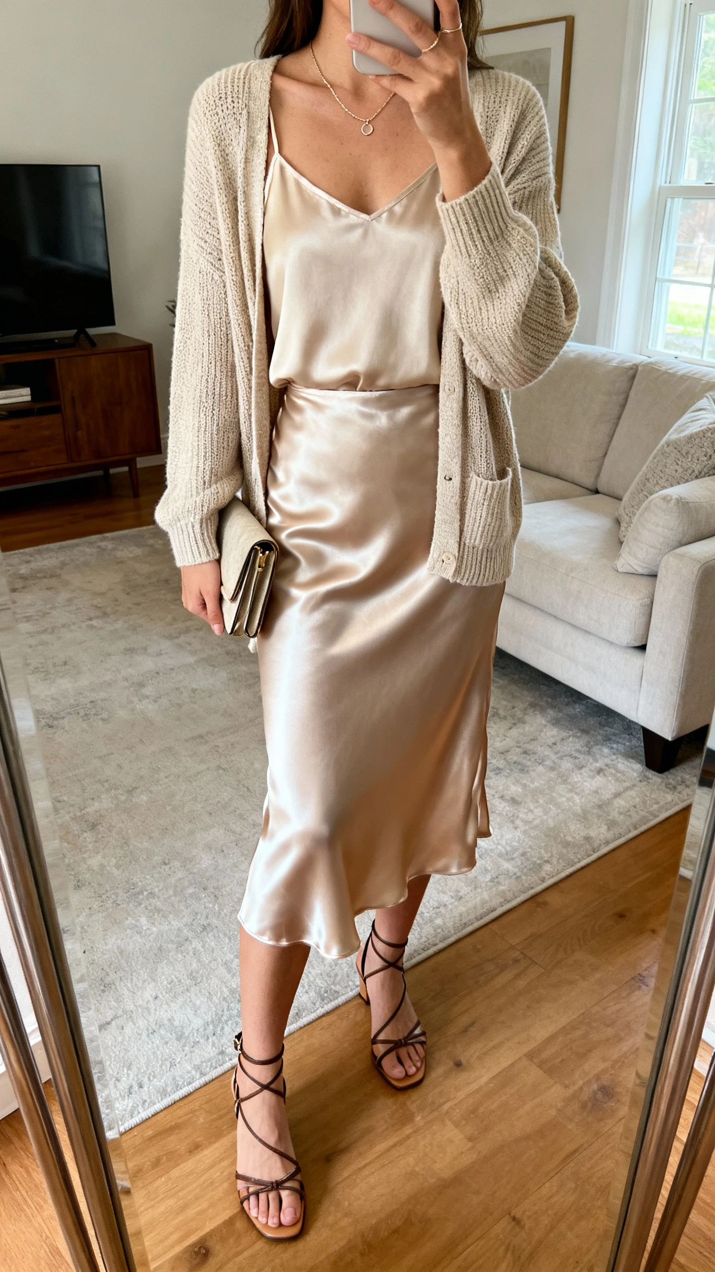 iPhone mirror selfie of a woman in a satin slip skirt with a matching knit cardigan set (camisole + cardigan), delicate necklace, strappy heels, and a clutch, face not visible, cropped at neck, living room mirror, soft indoor lighting, iPhone photo quality.
