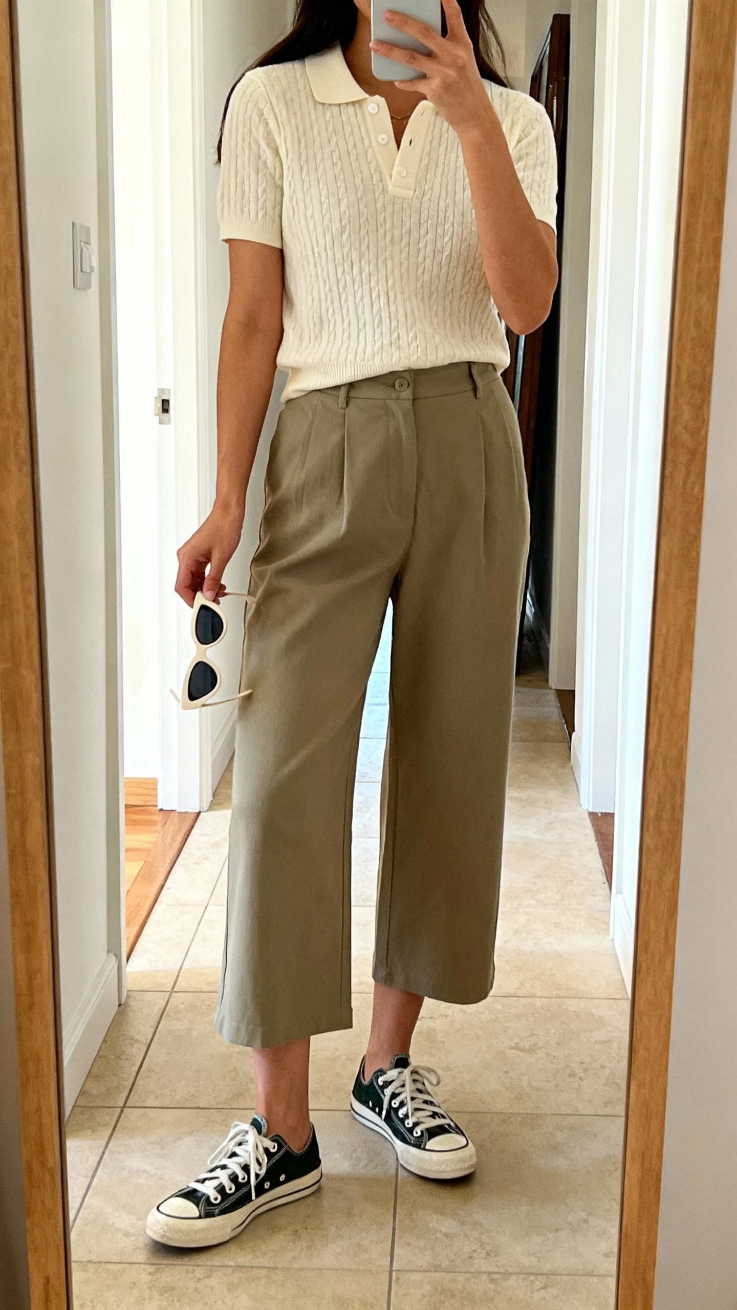 iPhone mirror selfie of a woman in a short-sleeve knit polo with cropped wide-leg pants, retro sneakers, cat-eye sunglasses in hand, face not visible, full-length mirror in entryway with natural daylight, iPhone photo quality.