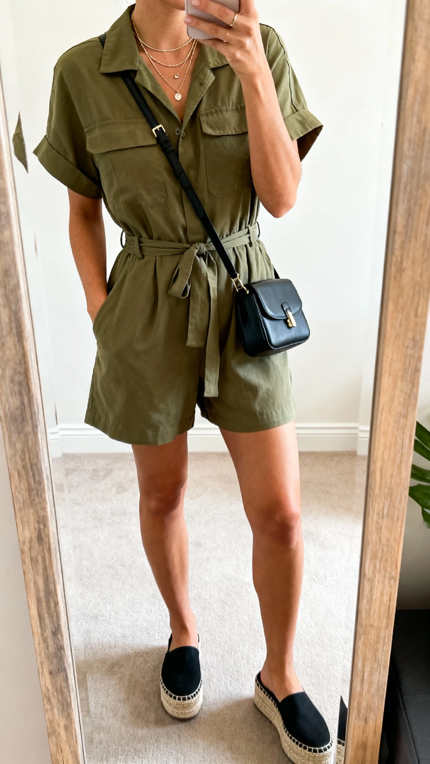 iPhone mirror selfie of a woman in a short-sleeve utility jumpsuit in olive with a cinched waist, layered delicate necklaces, black crossbody, platform espadrilles, face not visible, cropped at shoulders, full-length mirror, indoor daylight, iPhone photo quality.