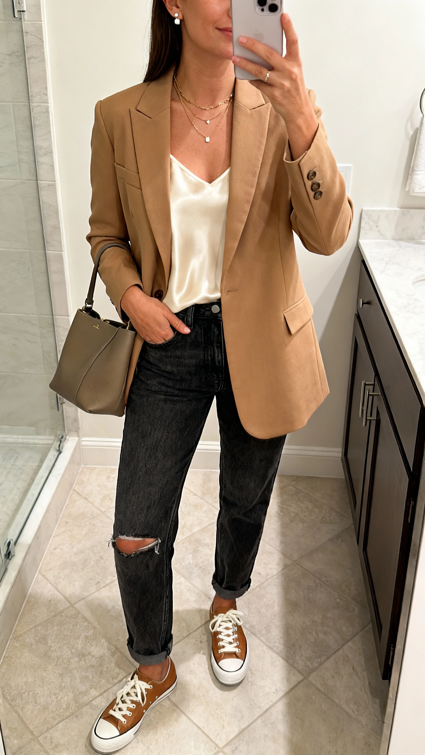 iPhone mirror selfie of a woman in a single-breasted camel blazer, dark-wash mom jeans (no rips), a silky cream camisole, lo-top leather sneakers, delicate layered necklaces and stud earrings, carrying a structured tote, face not visible, bathroom mirror, indoor morning light, iPhone photo quality.