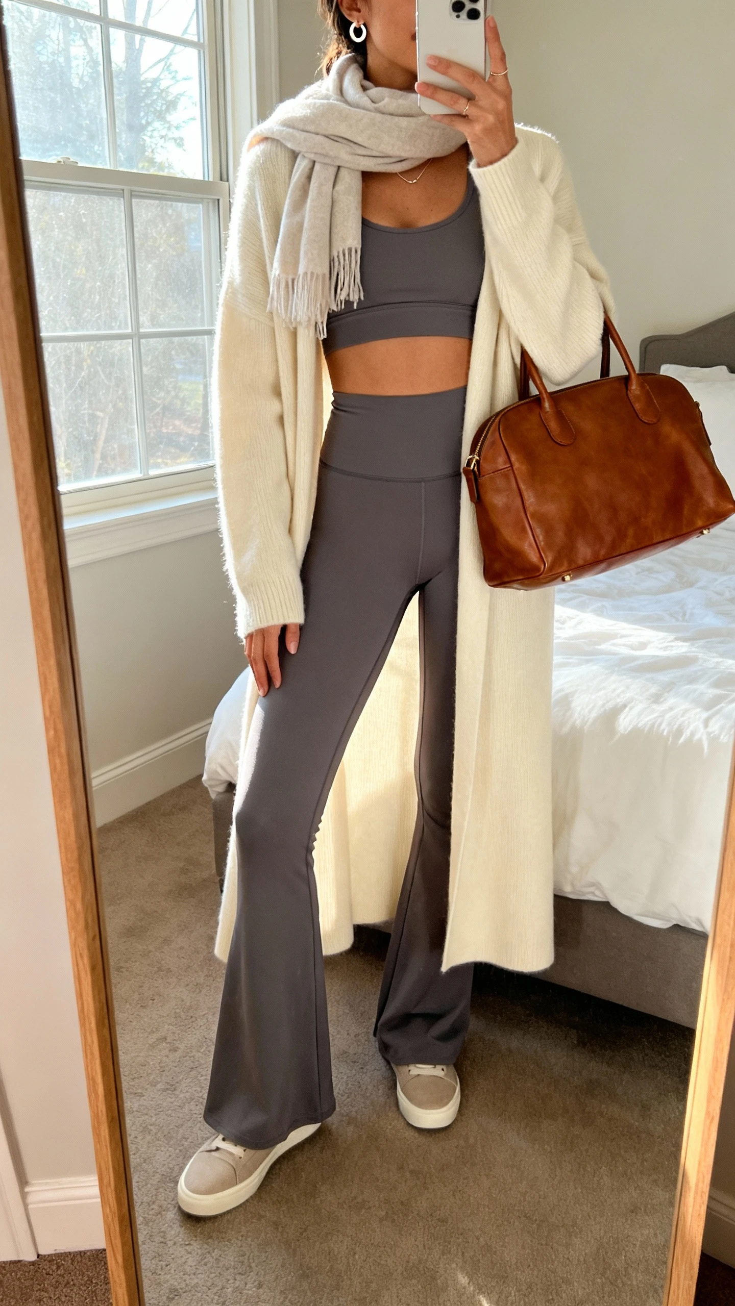 iPhone mirror selfie of a woman in a slate longline sports bra, flared leggings with a smoothing waistband, a cream knee-length cardigan coat, minimal slip-on sneakers, a soft scarf, delicate huggie earrings, and a leather-look tote; face not visible, cropped at neck, bedroom mirror, warm window light, iPhone photo quality.