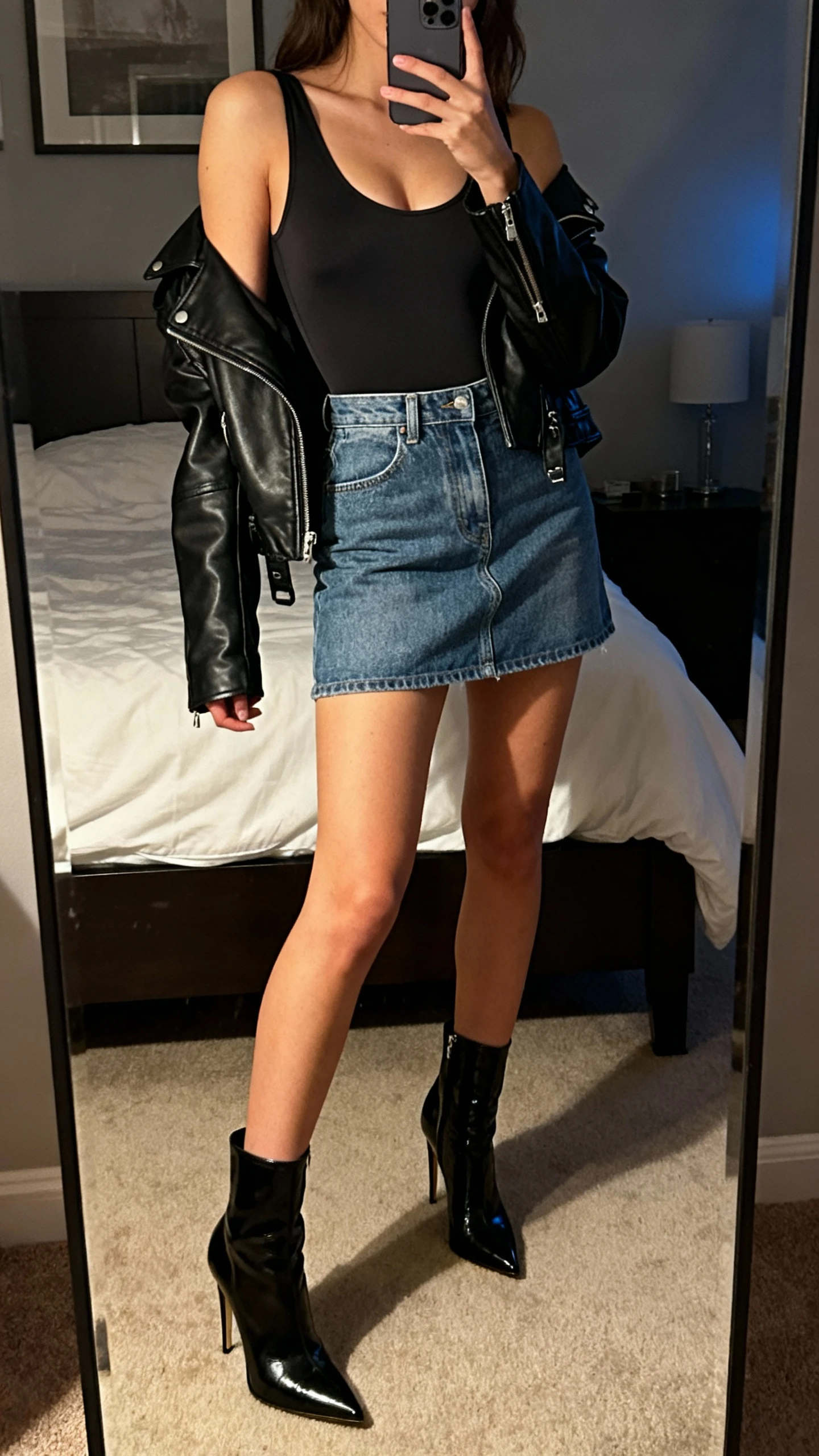 iPhone mirror selfie of a woman in a sleek bodysuit tucked into a denim mini skirt, moto jacket thrown over shoulders, and stiletto boots, face not visible, bedroom mirror, evening indoor light, iPhone photo quality.
