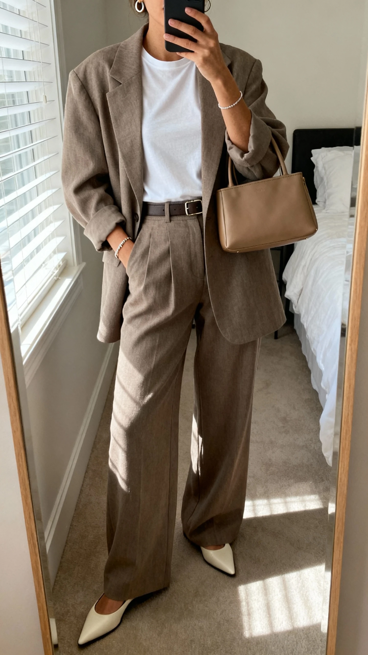 iPhone mirror selfie of a woman in a taupe oversized blazer with coordinating high-waist wide-leg trousers, white fitted crewneck tee, pointed slingback flats, minimal leather tote, slim belt, delicate bracelet, and small hoops, face not visible, cropped at shoulders, bedroom mirror with window light, iPhone photo quality.