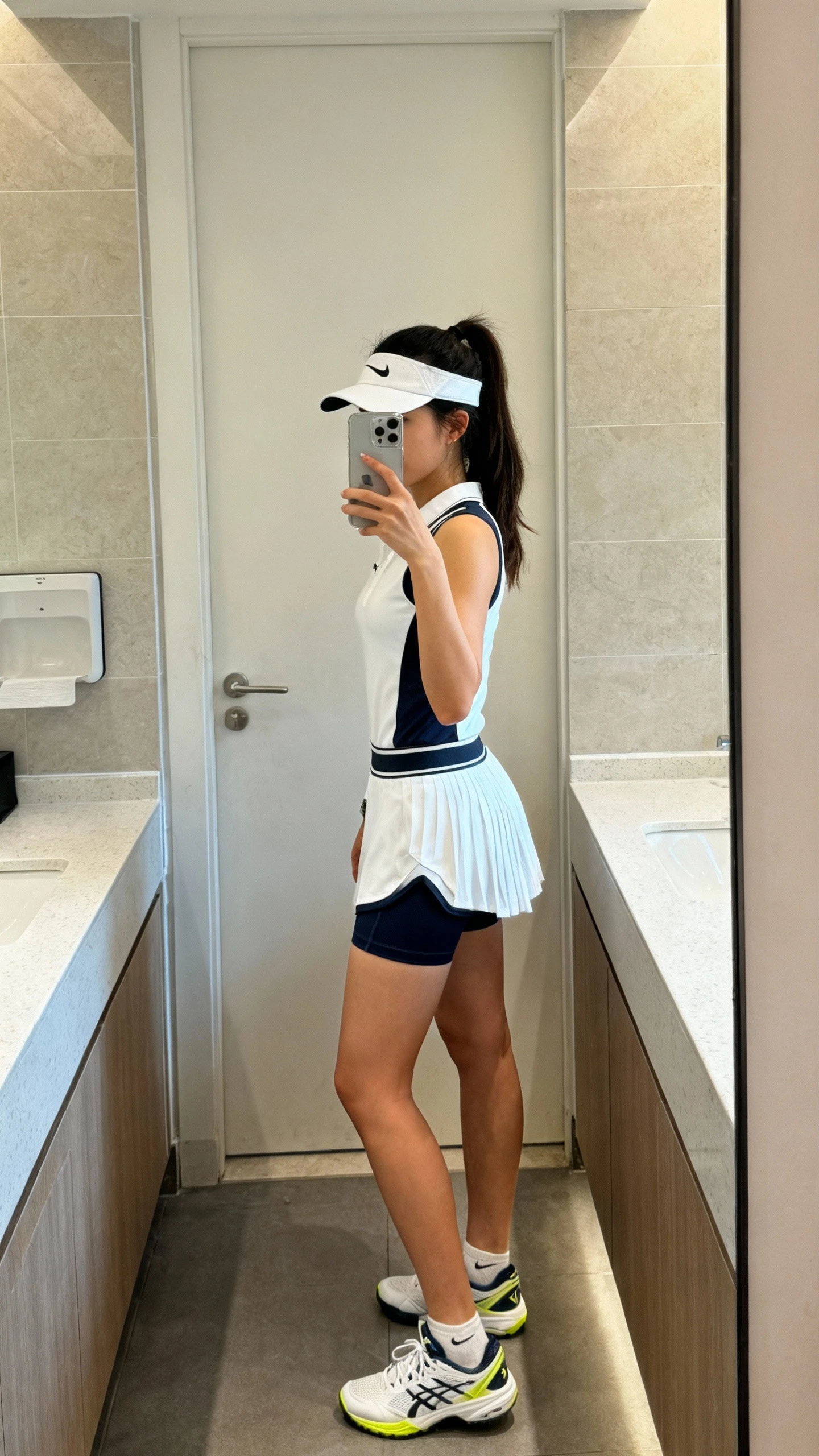 iPhone mirror selfie of a woman in a tennis dress with built-in shorts, face not visible, side angle in a bathroom mirror, visor and court sneakers, natural indoor lighting, iPhone photo quality.