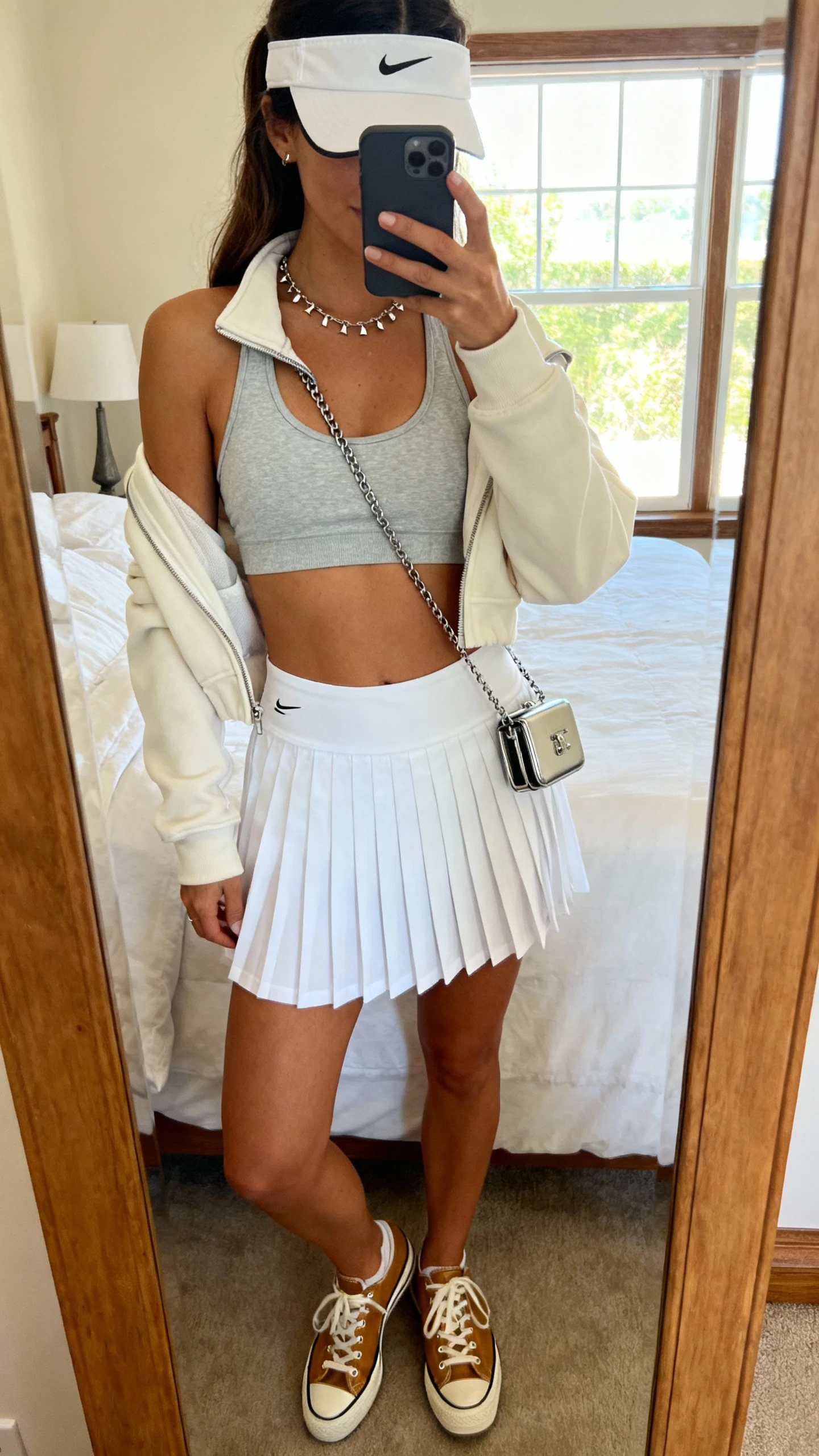 iPhone mirror selfie of a woman in a white pleated athletic skort, a light grey fitted racerback sports bra, a cropped half-zip, retro-inspired leather sneakers, a visor, delicate chain necklace with studs, and a compact crossbody bag; face not visible, cropped at shoulders, bedroom mirror, soft window light, iPhone photo quality.