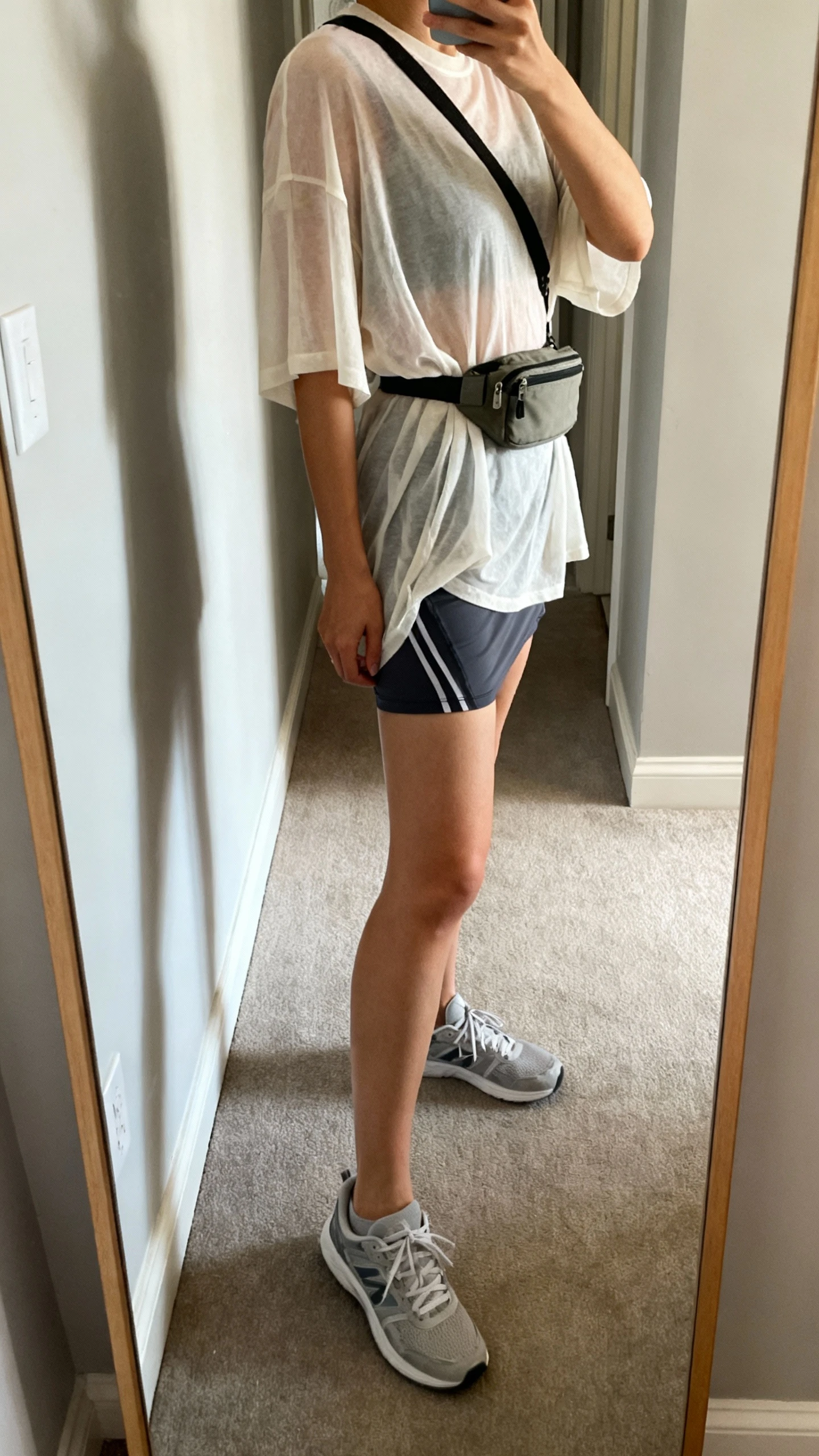 iPhone mirror selfie of a woman in an airy oversized tee and a lined running skirt, face not visible, side angle in a hallway mirror, casual sneakers and a small crossbody belt bag, soft morning light, iPhone photo quality.
