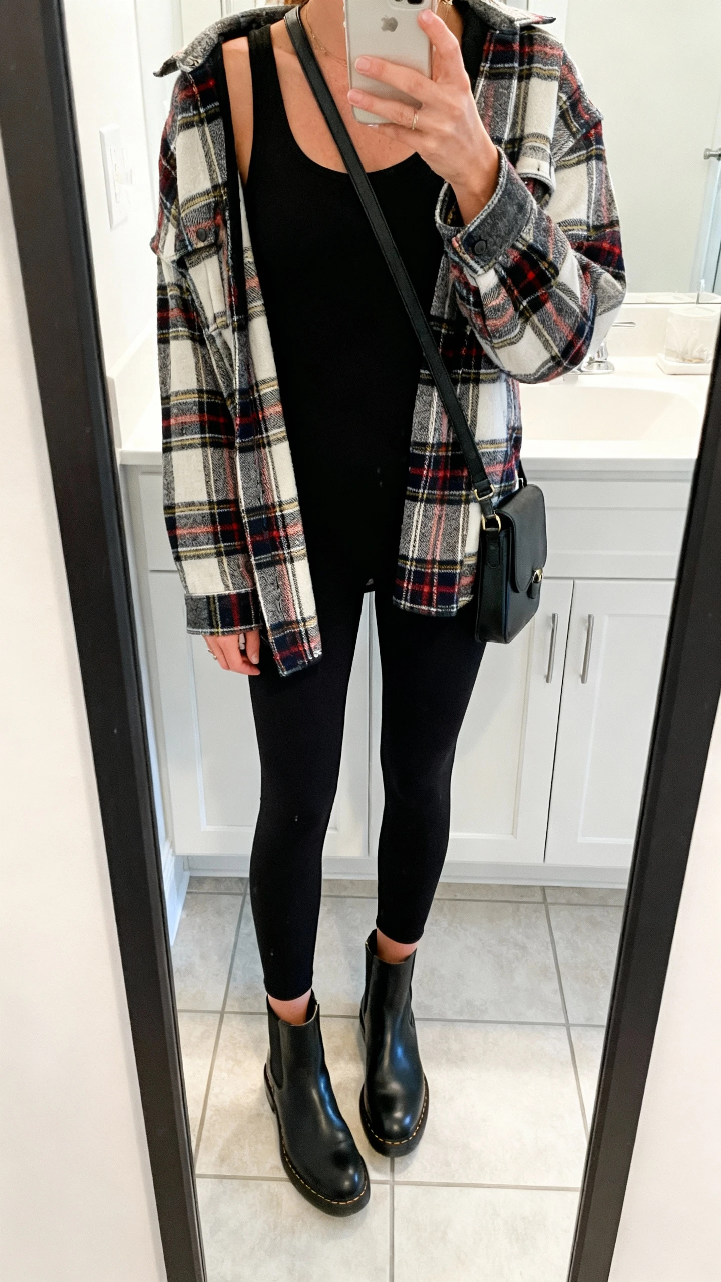 iPhone mirror selfie of a woman in an all-black base: black leggings and black longline tank, topped with a plaid statement shacket, black Chelsea boots, and black crossbody, face not visible, cropped at shoulders, bathroom mirror, bright indoor light, iPhone photo quality.