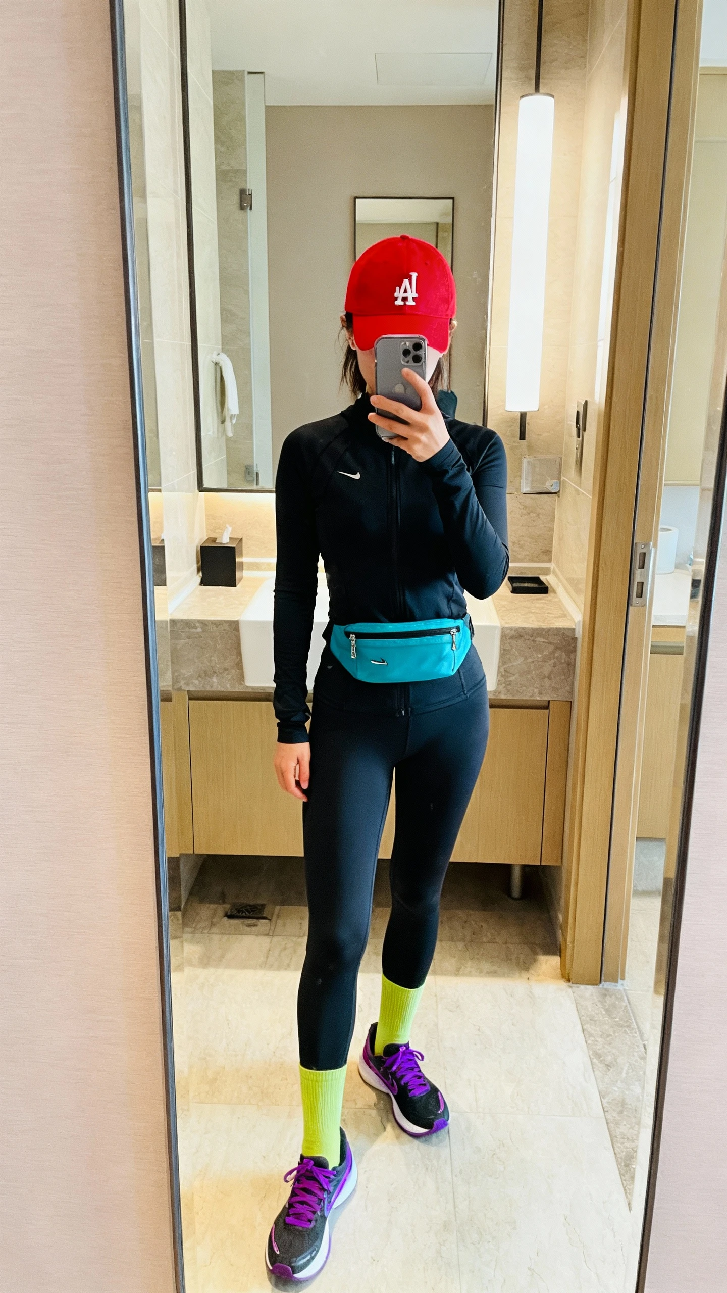 iPhone mirror selfie of a woman in an all-black gym set with loud color-blocked accessories: red baseball cap, cobalt waist pack, chartreuse socks, and purple-accent running shoes, face not visible, cropped at neck, bathroom mirror, bright indoor lighting, iPhone photo quality.