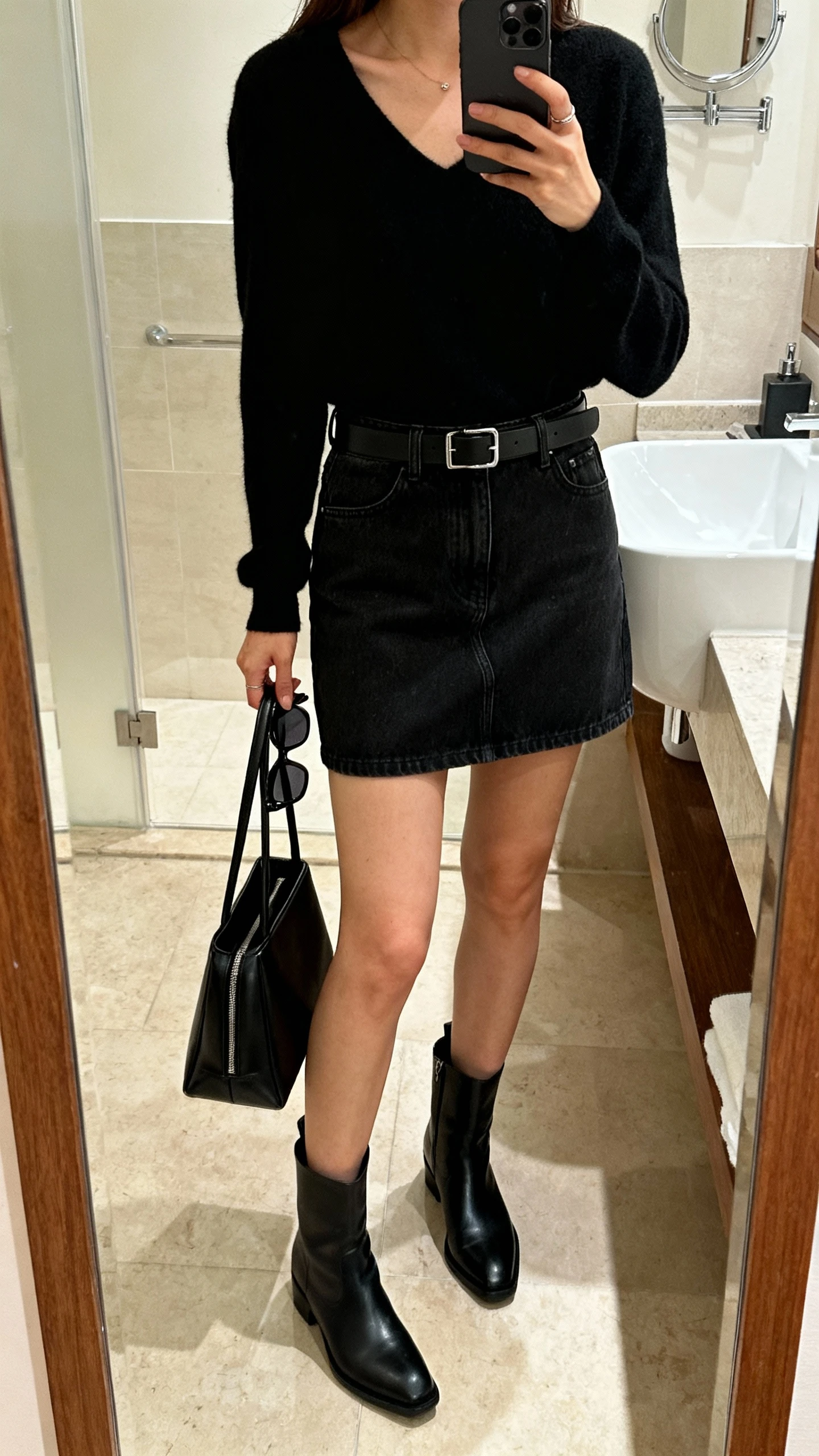 iPhone mirror selfie of a woman in an all-black look: black denim mini skirt, black top, black belt, sleek black ankle boots, sharp sunglasses in hand, black structured bag, face not visible, cropped at neck, bathroom mirror, indoor lighting, iPhone photo quality.