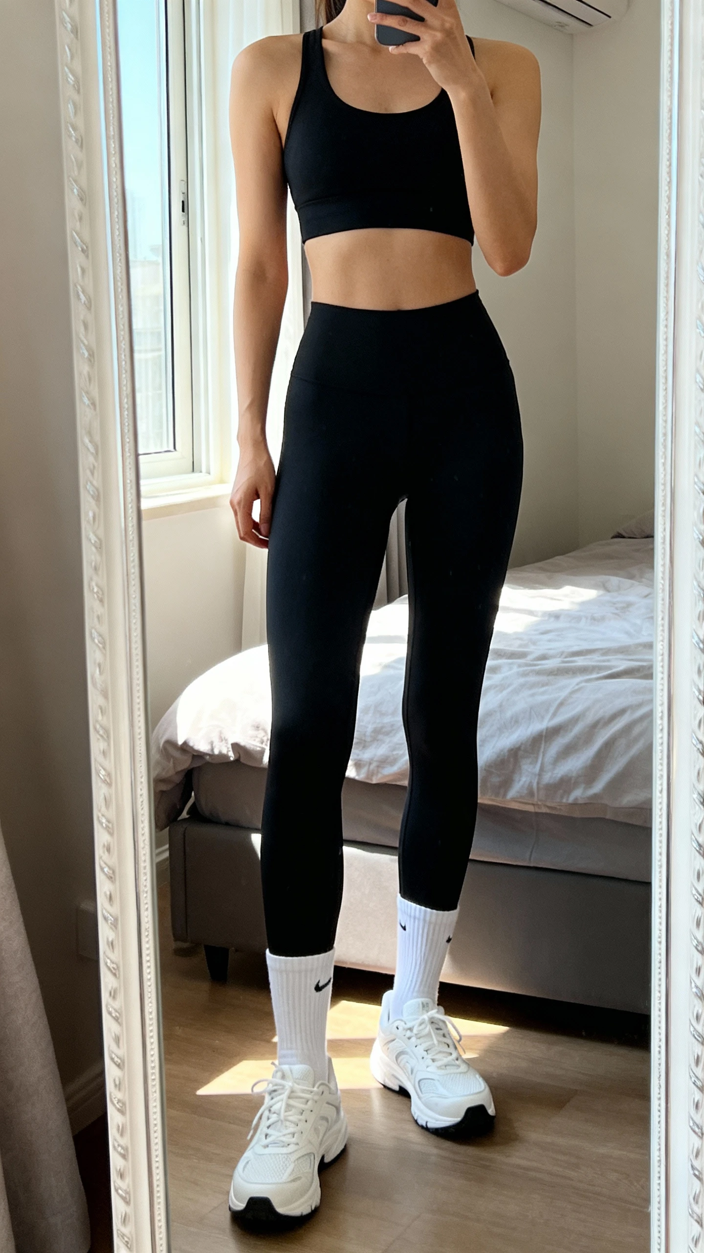 iPhone mirror selfie of a woman in an all-black set (black compression leggings and black sports bra) with white crew socks and white training shoes, face not visible, cropped at shoulders, full-length mirror in bedroom, soft afternoon window light, iPhone photo quality.