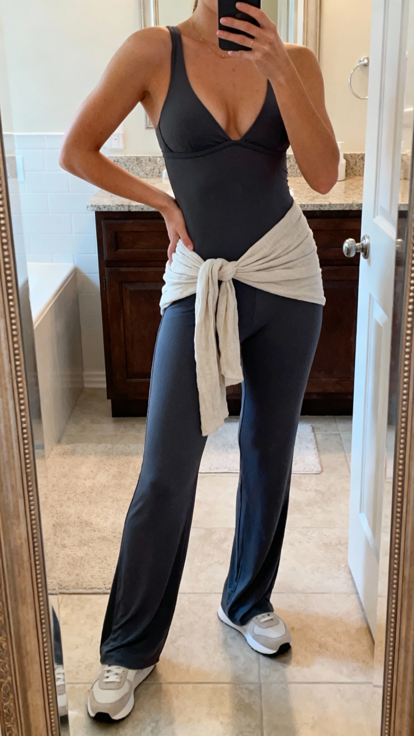 iPhone mirror selfie of a woman in an all-day one-piece jumpsuit with built-in bra, sleek ankle-length cut, minimalist trainers, and a light wrap tied around hips, face not visible, bathroom mirror, soft indoor lighting, iPhone photo quality.
