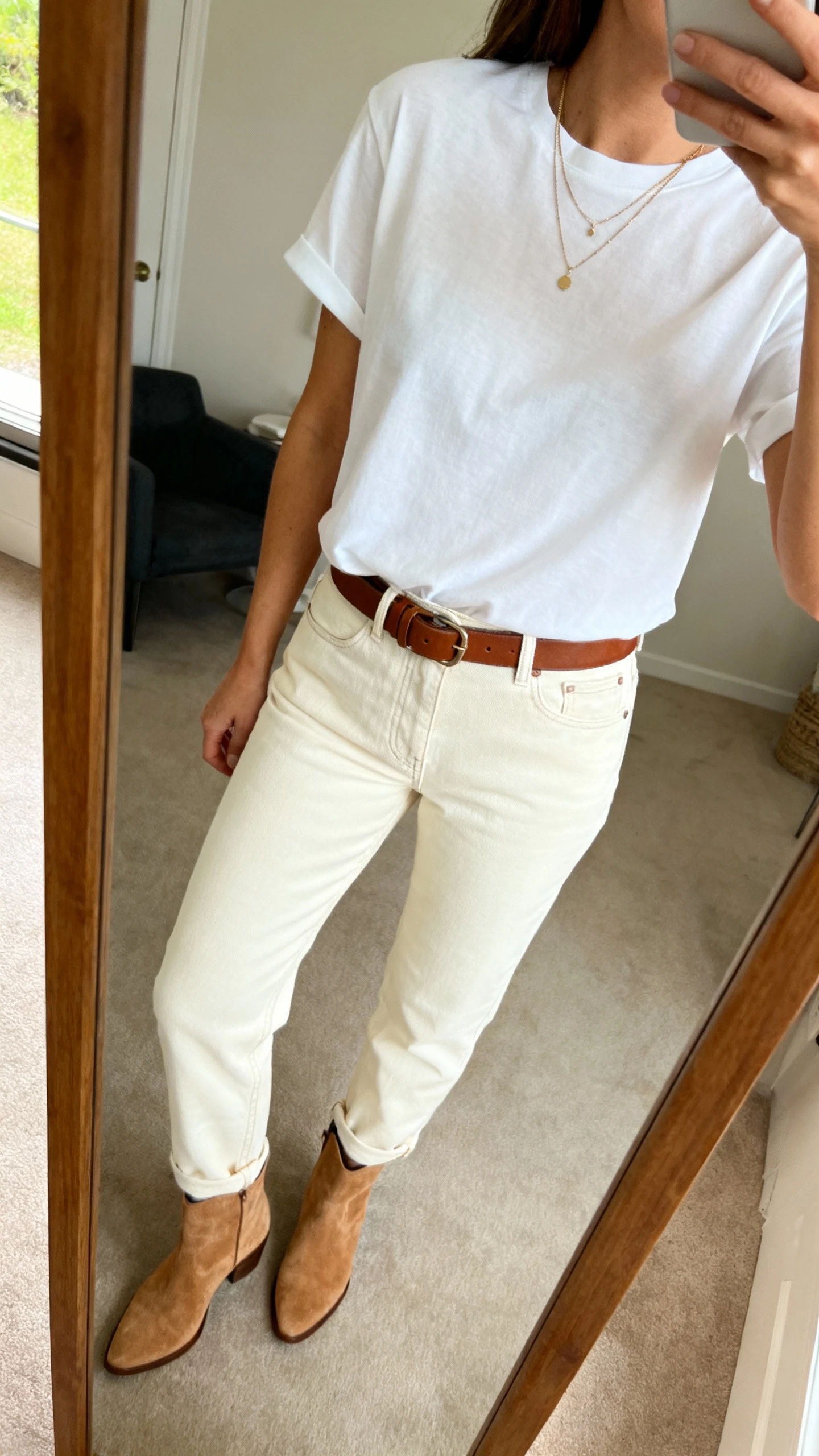 iPhone mirror selfie of a woman in an elevated white tee tucked into cream straight-leg denim, brown leather belt, camel suede ankle boots, delicate layered necklace, face not visible, side angle crop at shoulders, full-length mirror, indoor natural light, iPhone photo quality.
