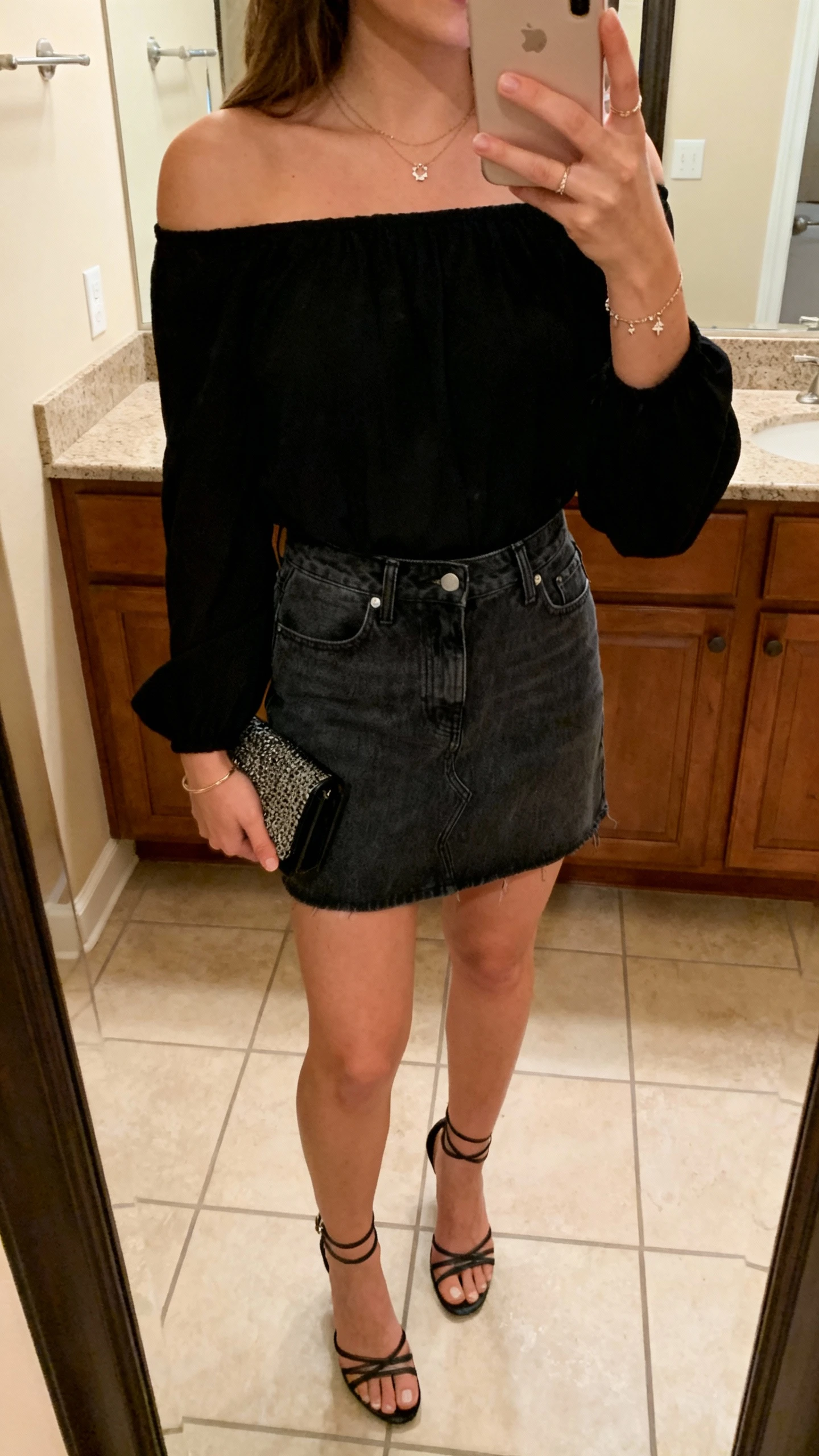 iPhone mirror selfie of a woman in an off-shoulder black top, dark-wash denim mini skirt, strappy heels, and a small clutch with delicate jewelry, face not visible, cropped at shoulders, bathroom mirror, warm indoor lighting, iPhone photo quality.