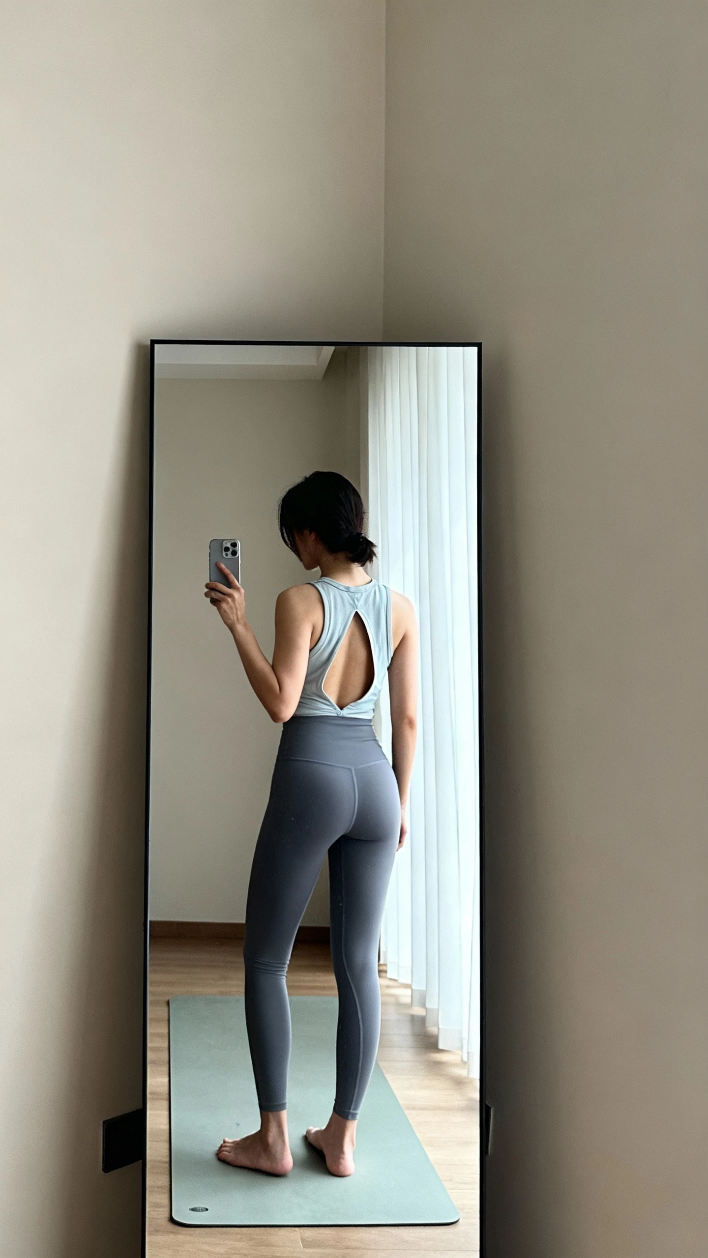 iPhone mirror selfie of a woman in an open-back tank and seamless leggings, face not visible, cropped at shoulders to show open back, studio corner mirror with calm natural light, barefoot with a simple yoga mat, iPhone photo quality.