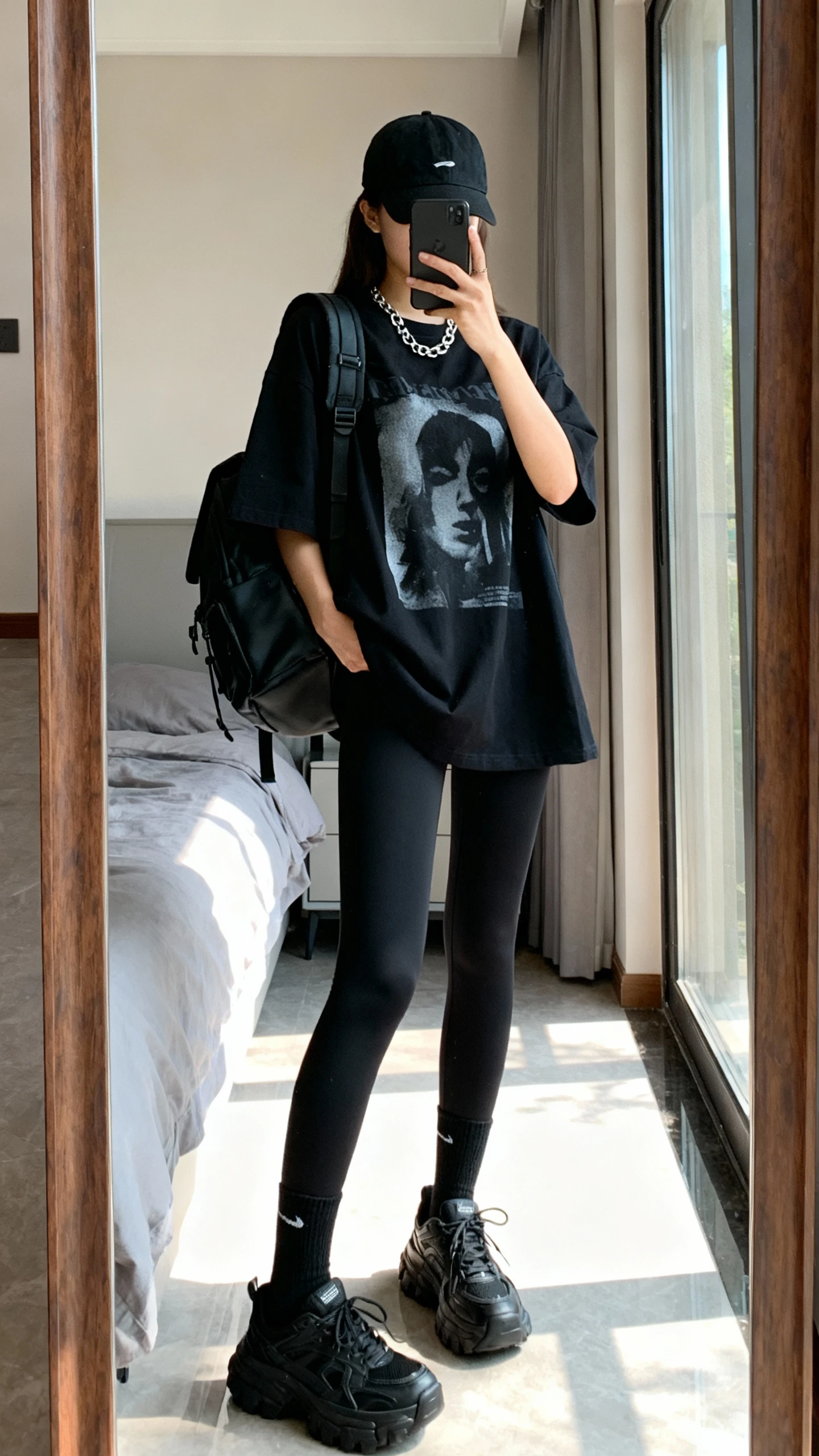 iPhone mirror selfie of a woman in an oversized black graphic tee, black compressive leggings, chunky black trainers, black crew socks, black baseball cap, structured black backpack, and a simple silver chain necklace, face not visible, bedroom full-length mirror, afternoon window light, iPhone photo quality.
