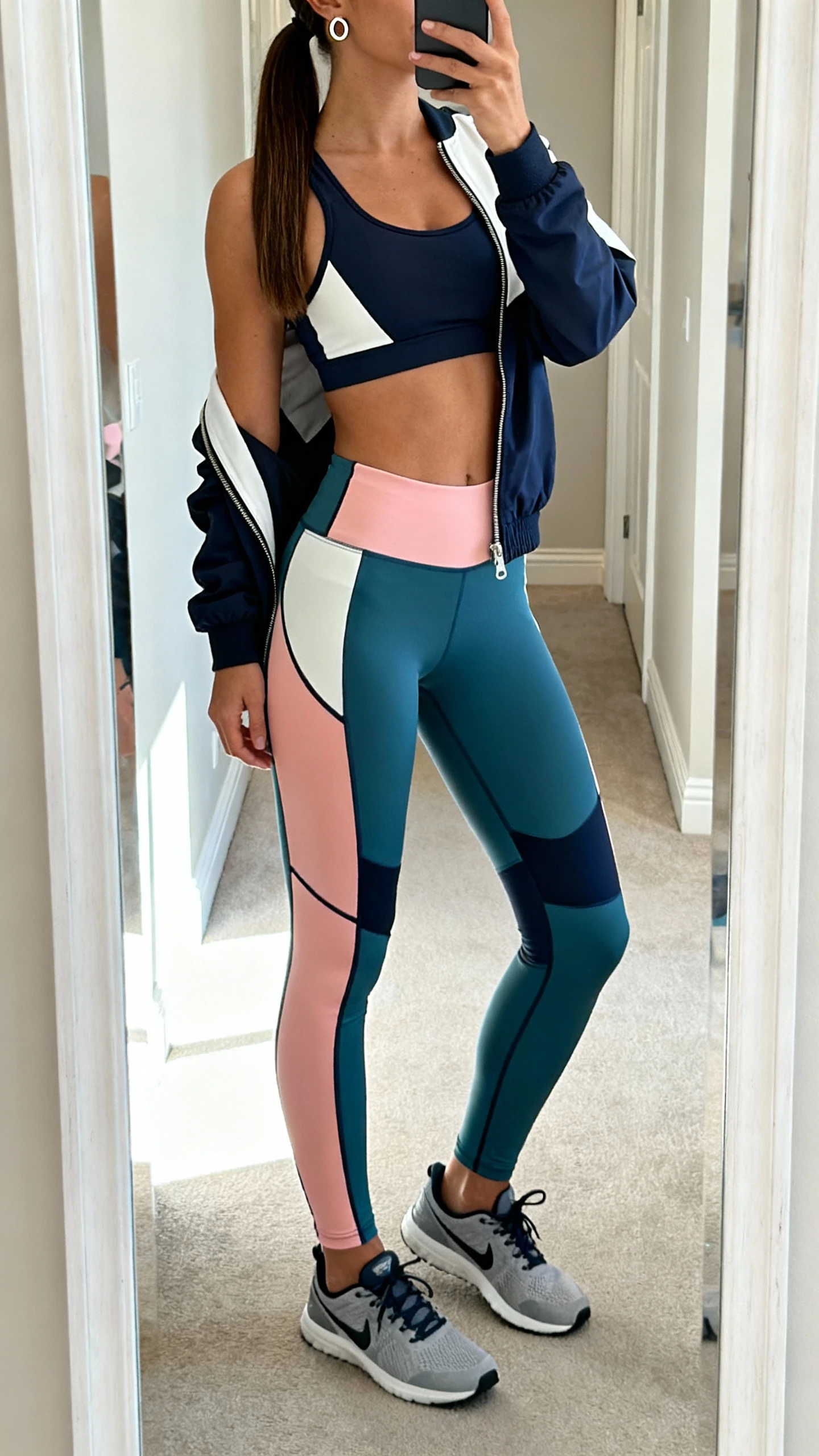 iPhone mirror selfie of a woman in color-blocked leggings with strategic side panels, a high-support sports bra, breathable training sneakers, lightweight zip-front track jacket slightly unzipped, simple huggie earrings and sleek ponytail, face not visible, cropped at neck, hallway full-length mirror with bright daylight, iPhone photo quality