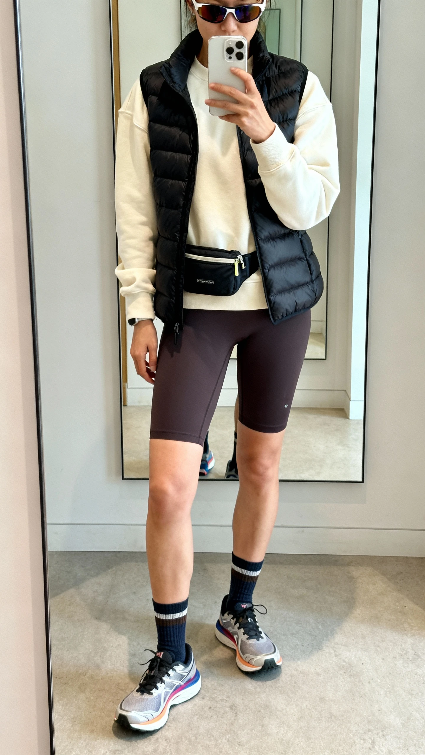 iPhone mirror selfie of a woman in deep charcoal 7-inch contour bike shorts, a cream midweight crewneck sweatshirt, a black streamlined quilted vest, performance running shoes with subtle color accents, athletic ankle socks, a hands-free belt bag, and sport sunglasses; face not visible, cropped at shoulders, full-length mirror, natural daylight, iPhone photo quality.