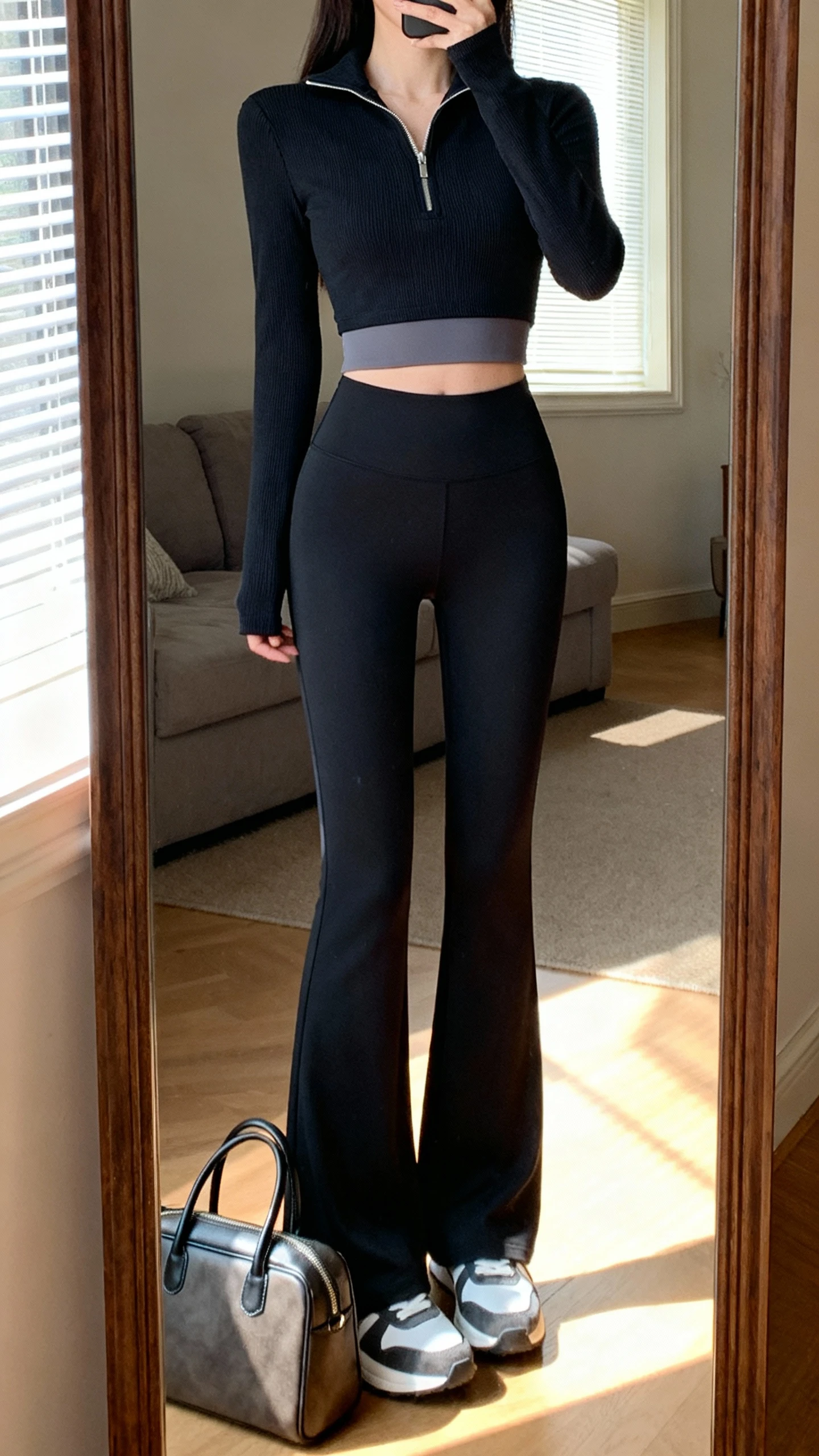 iPhone mirror selfie of a woman in high-waisted flared leggings, a slim half-zip pullover with a seamless crop underlayer peeking, clean low-profile sneakers, structured tote by her feet, face not visible, cropped at neck, living room full-length mirror with morning window light, iPhone photo quality